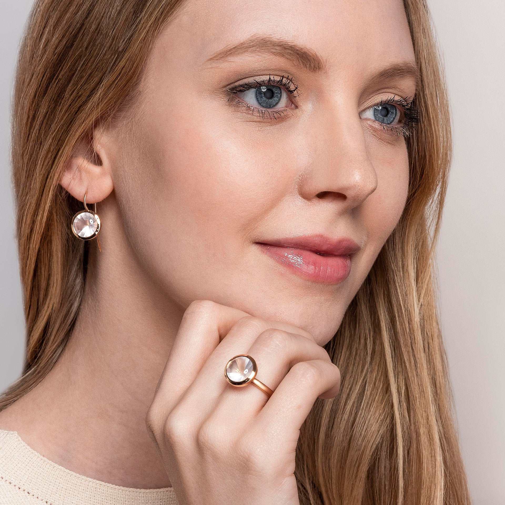 Eine Frau mit langem blondem Haar und leichtem Make-up lächelt sanft. Sie trägt den Schullin ROSENQUARZ RING BUFFTOP - einen goldenen Ring, der mit einem runden, klaren Edelstein besetzt ist - und dazu passende goldene Ohrringe, während sie ihre Hand neben ihr Gesicht legt.