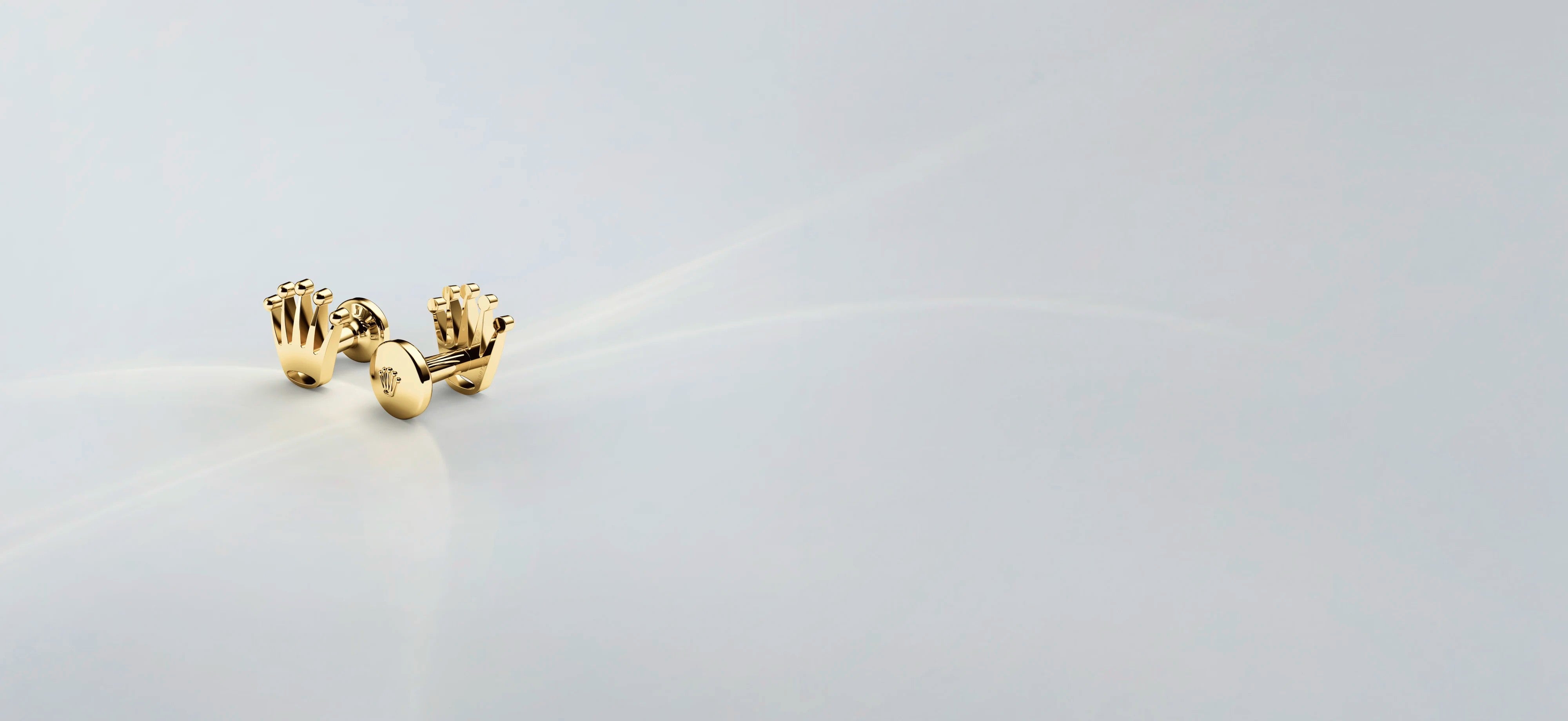 Ein Paar goldene Manschettenknöpfe in Form von Händen auf einer glatten, hellgrauen Oberfläche mit sanfter Beleuchtung und viel freiem Raum um sie herum.