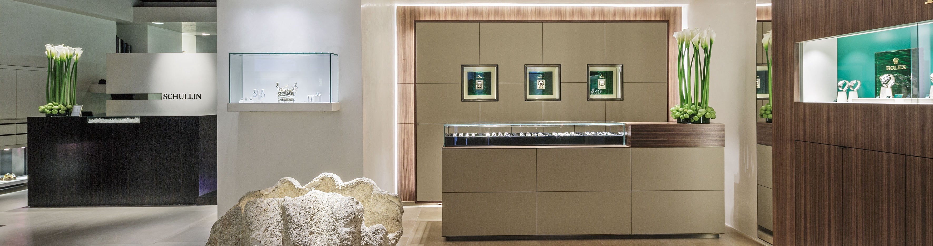 Ein modernes Interieur eines Luxusuhrengeschäfts mit eleganter Auslage und Beleuchtung. In Vitrinen werden Uhren ausgestellt, links steht ein dunkler Tresen mit der Aufschrift „SCHILLIN“. Überall im Raum sind dekorative Blumenarrangements platziert, die für eine elegante Note sorgen.