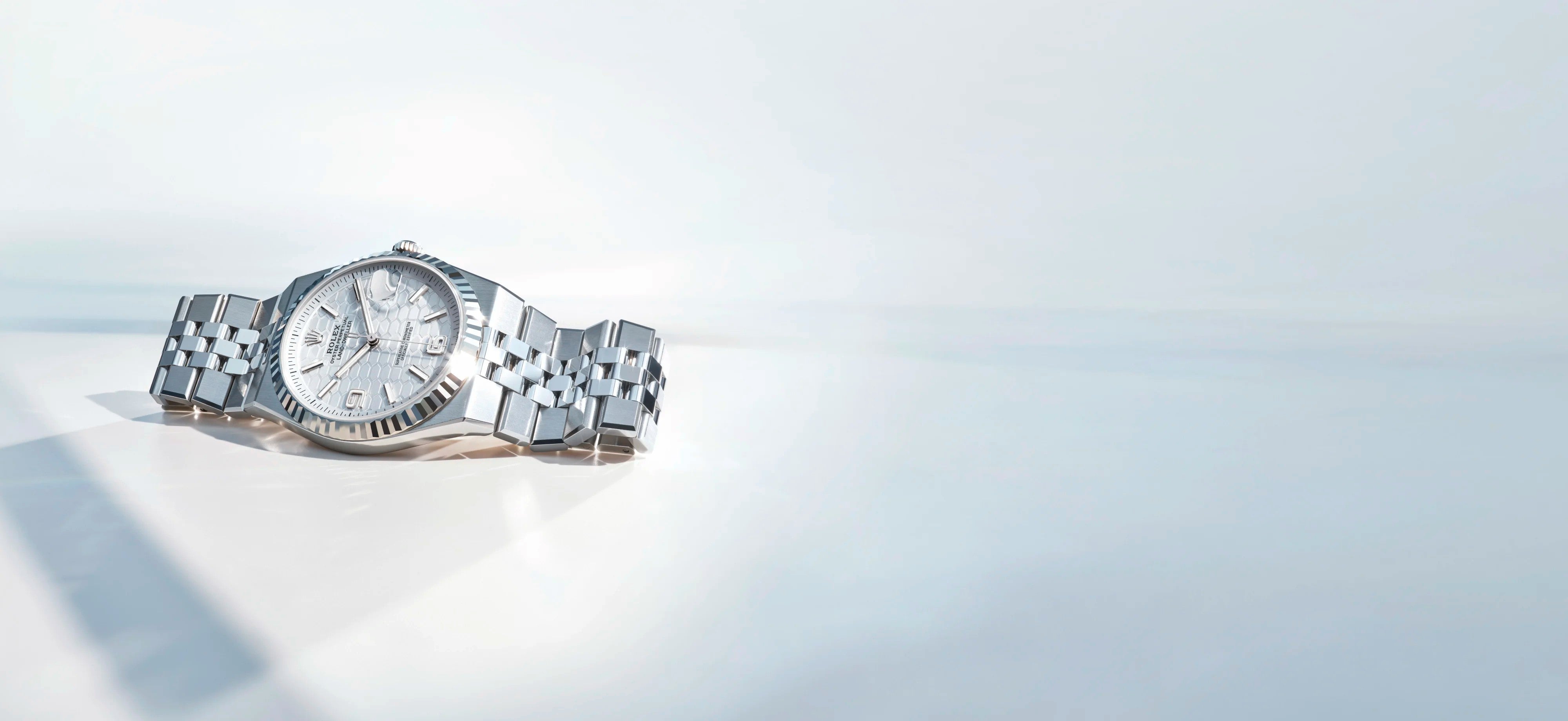 Eine silberne Luxusarmbanduhr mit Metallgliederband liegt auf einer glänzenden weißen Oberfläche, die das Licht reflektiert. Das Ziffernblatt zeigt die Uhrzeit und das Datum an, und das Gesamtbild wirkt sauber und elegant.