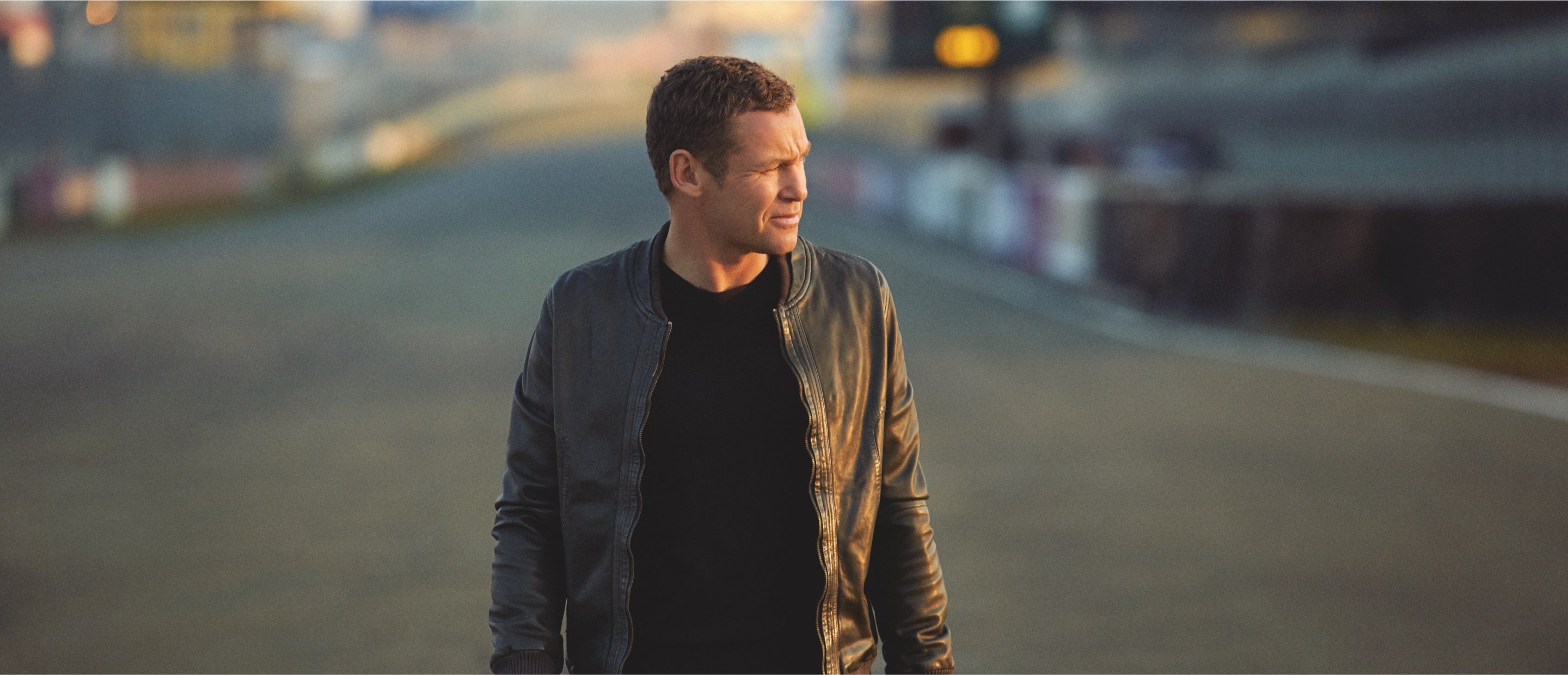 Eine Person in einer Lederjacke läuft auf einer Rennstrecke mit verschwommenem Hintergrund. Die Beleuchtung lässt vermuten, dass es Morgen- oder Abenddämmerung ist, und wirft einen warmen Schein auf die Szene.