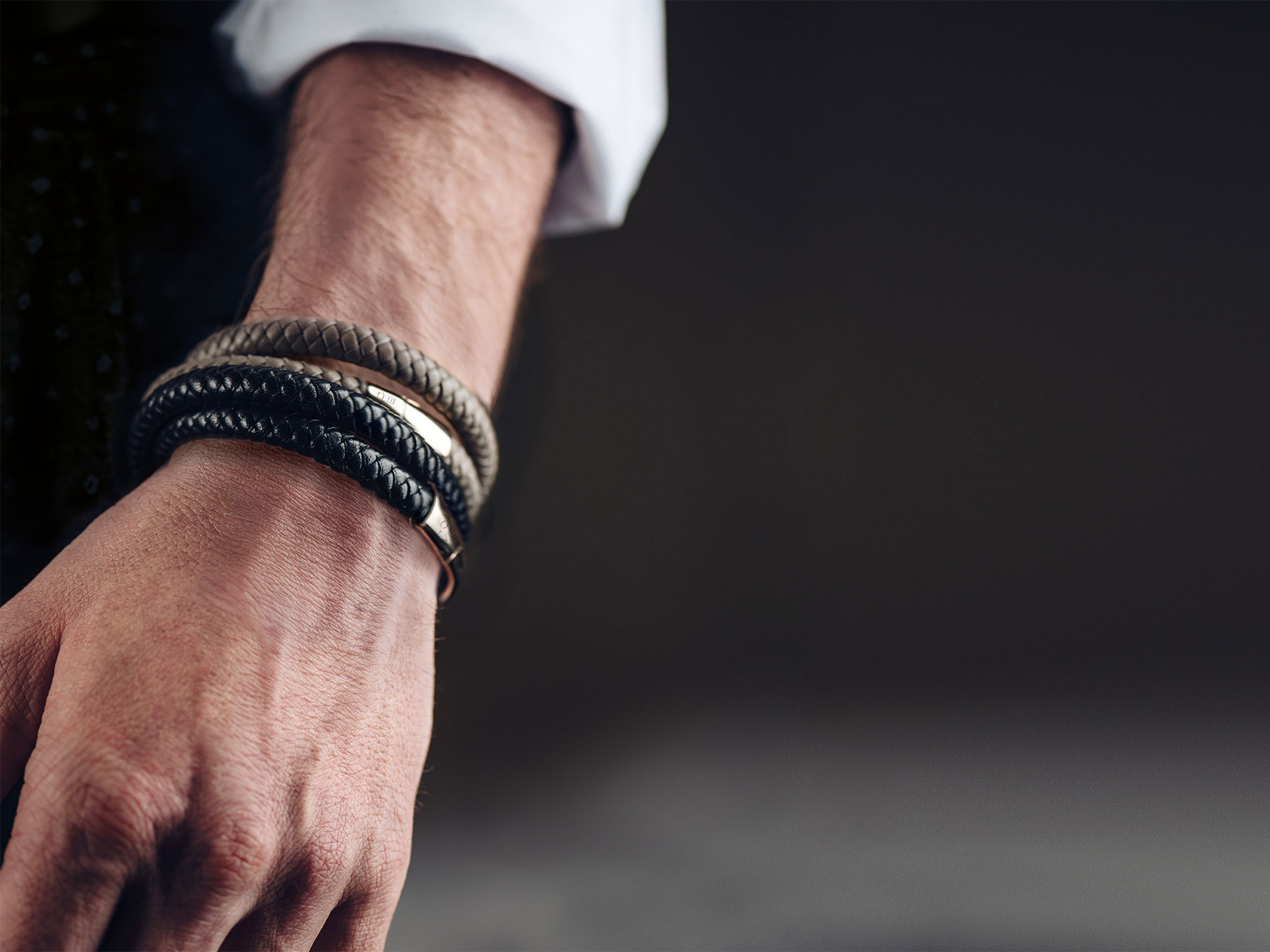 Eine Nahaufnahme zeigt ein Handgelenk mit drei Schullin POWER ARMBAND MIT GRAVUR Lederarmbändern in Schwarz, Braun und Beige mit metallischen Akzenten, getragen über einem weißen langärmeligen Hemd vor einem dunklen, verschwommenen Hintergrund.