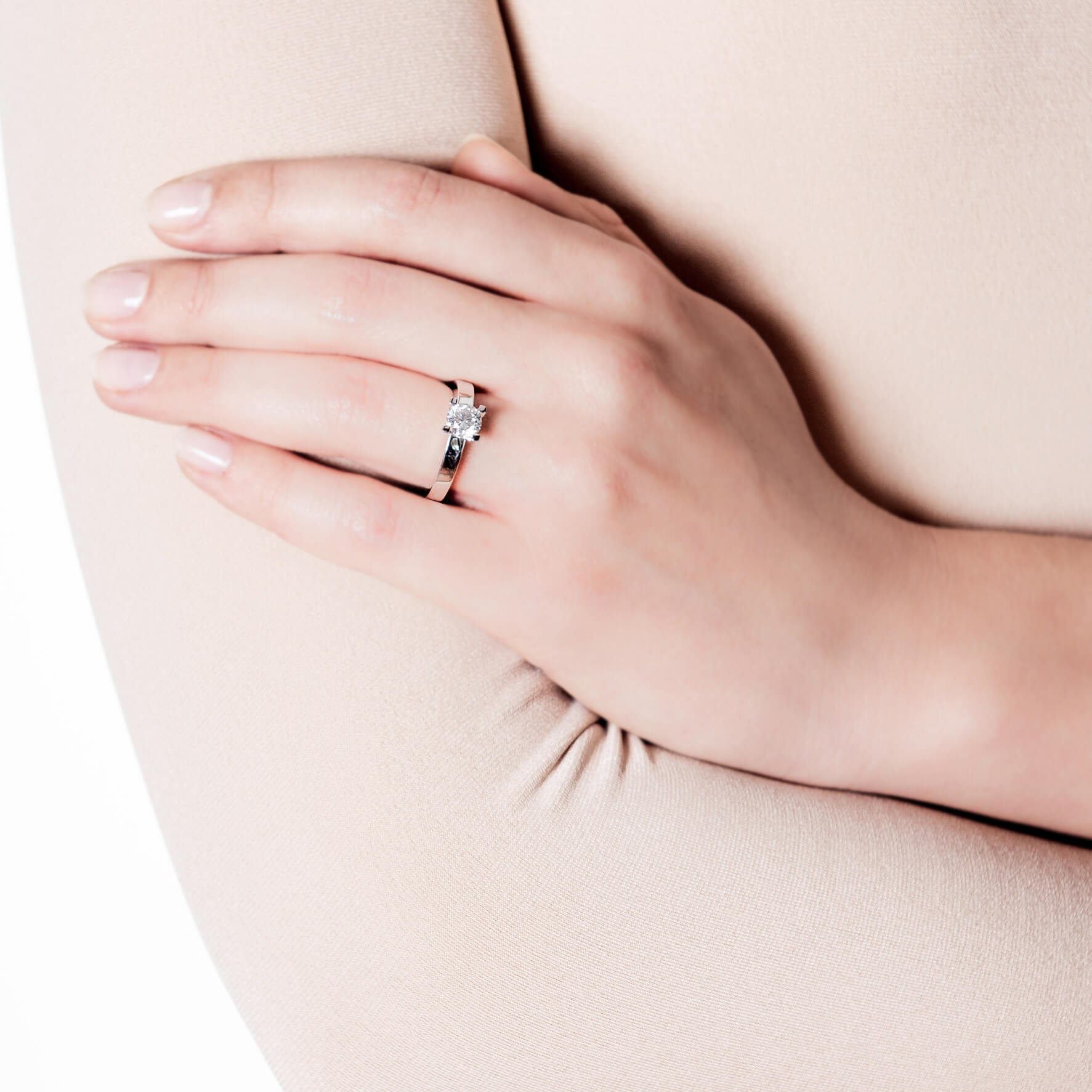 Eine Nahaufnahme einer Hand, die auf beigem Stoff ruht und mit dem Schullin VICTORY SOLITÄR 0,71 CARAT Weißgold-Verlobungsring mit einem markanten ovalen Edelstein am Ringfinger geschmückt ist. Die Fingernägel sind mit einem leichten, natürlichen Lack manikürt.