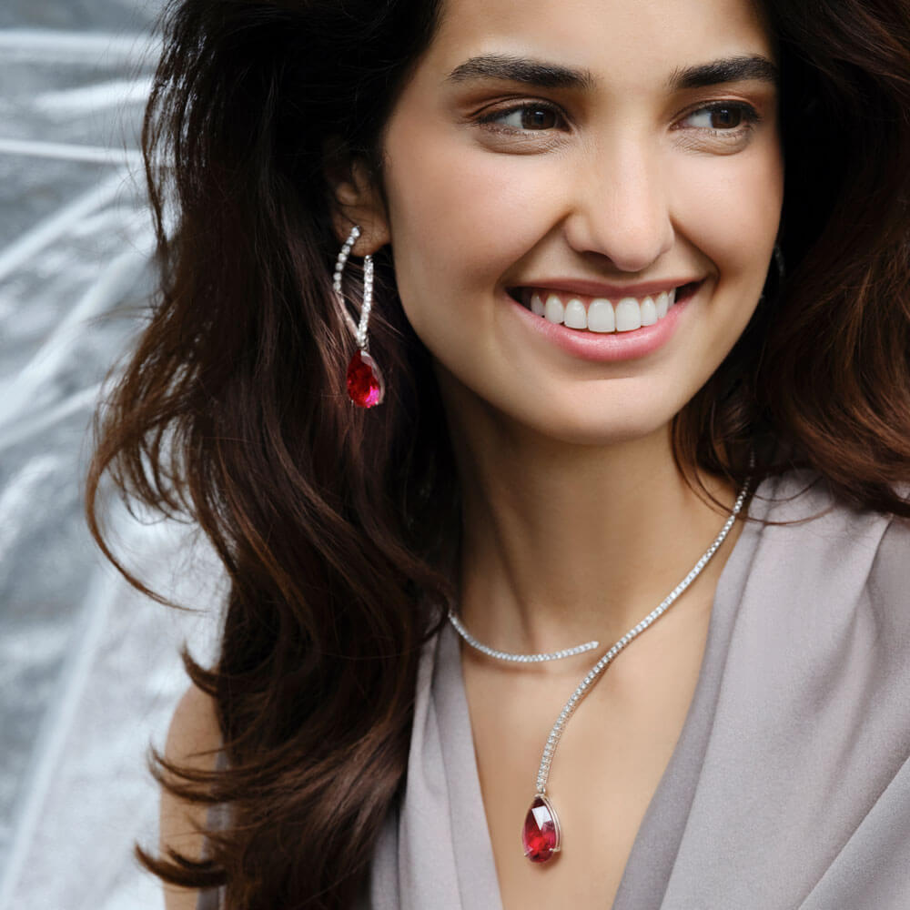 Eine Frau mit langen dunklen Haaren lächelt und trägt eleganten, funkelnden Schmuck. Sie trägt Schullins DIAMANT-OHRRINGE mit Rubellit-Tropfen und eine passende Halskette aus Weißgold. Der sanft verschwommene Hintergrund unterstreicht ihre anmutige Pose und die stilvollen Accessoires.