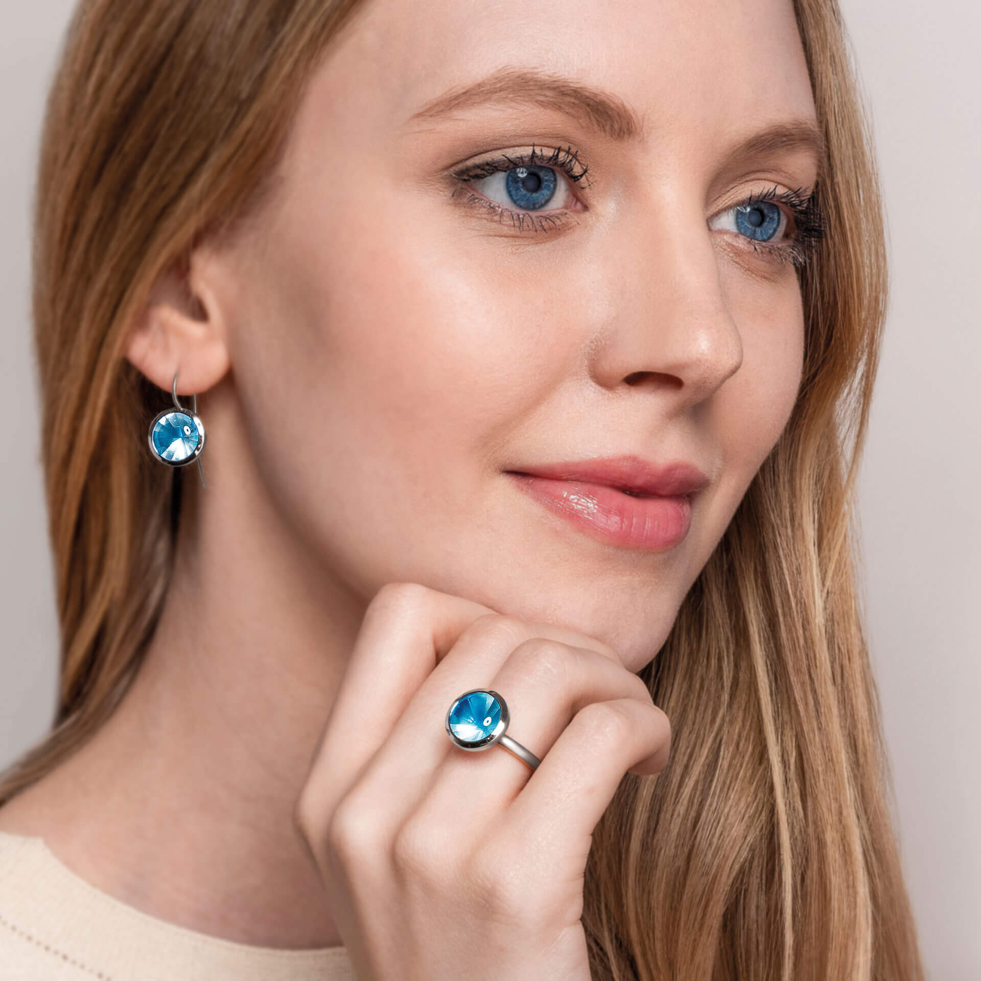 Eine Frau mit langen blonden Haaren trägt Schullin TOPAS OHRRINGE BUFFTOP Ohrringe in Weißgold und einen dazu passenden Ring. Sie lächelt leicht und blickt zur Seite, vor einem Hintergrund in sanften, neutralen Tönen.