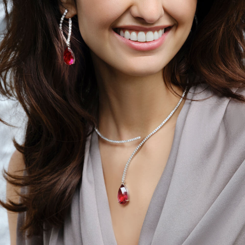 Eine Frau lächelt und trägt die Schullin DIAMANT HALSREIF-Halskette mit einem rubellitroten Anhänger und dazu passende Ohrringe. Ihr dunkles Haar fällt über ihre Schultern und passt perfekt zu ihrem hellgrauen Oberteil. Der sanft verschwommene Hintergrund betont die Eleganz ihres Ensembles.
