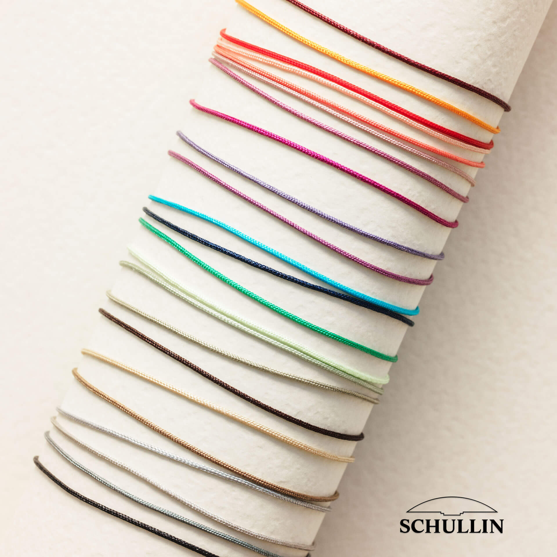 Eine Auswahl lebendiger INFINITY ARMBAND-Armbänder mit dünnen Kordeln und Infinity-Anhängern, elegant angeordnet auf einem zylindrischen beigen Ständer. Diese Armbänder sind aus Stoffband in verschiedenen Farben wie Pink, Orange und Rot gefertigt und strahlen Charme aus. In der unteren rechten Ecke ist das Logo der Marke Schullin zu sehen.