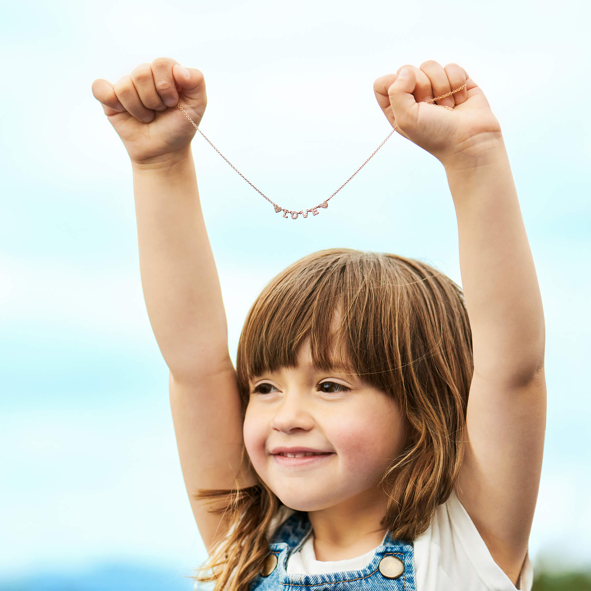 Ein kleines Kind mit langen braunen Haaren lächelt fröhlich, während es eine Schullin LOVE HALSKETTE aus Roségold hochhält und mit beiden Händen „Love“ buchstabiert. Der Himmel im Hintergrund ist hell und klar und hebt das Funkeln der Kette als perfektes Muttertagsgeschenk hervor.