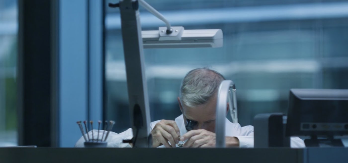 Eine Person mit grauem Haar sitzt an einem Schreibtisch und schaut durch ein Mikroskop. Die Büroeinrichtung umfasst einen Computermonitor und eine verstellbare Lampe. Große Fenster im Hintergrund zeigen eine verschwommene Ansicht eines Gebäudes.