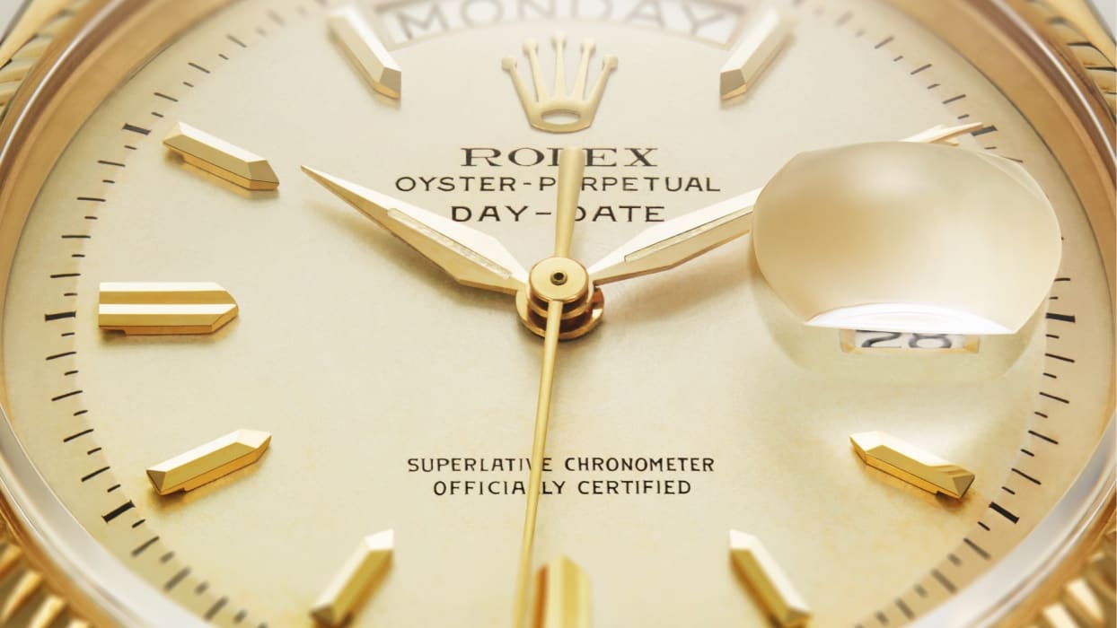 Nahaufnahme eines goldenen Zifferblatts einer Rolex Oyster Perpetual Day-Date-Uhr. Der Tag wird oben angezeigt und das Datum wird rechts vergrößert. Die Uhr verfügt über goldene Stundenmarkierungen und Zeiger sowie eine geriffelte Lünette, die das Zifferblatt umgibt.