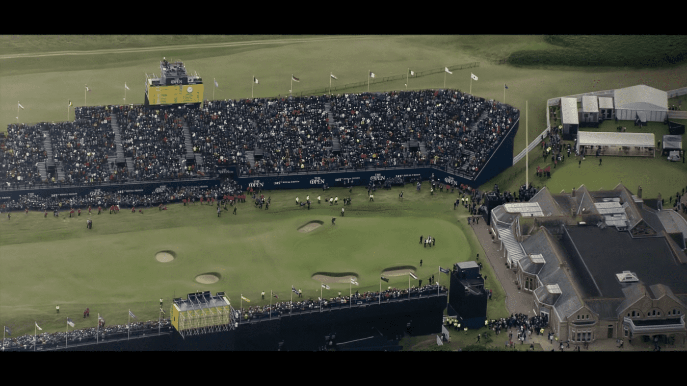 Luftaufnahme eines überfüllten Golfplatzes mit einer großen Zuschauertribüne und einem Clubhaus. Golfer befinden sich auf einem Grün mit Sandbunkern in der Nähe. Der Platz ist von üppigem Gras umgeben und vereinzelt sind Menschen, die das Geschehen verfolgen.