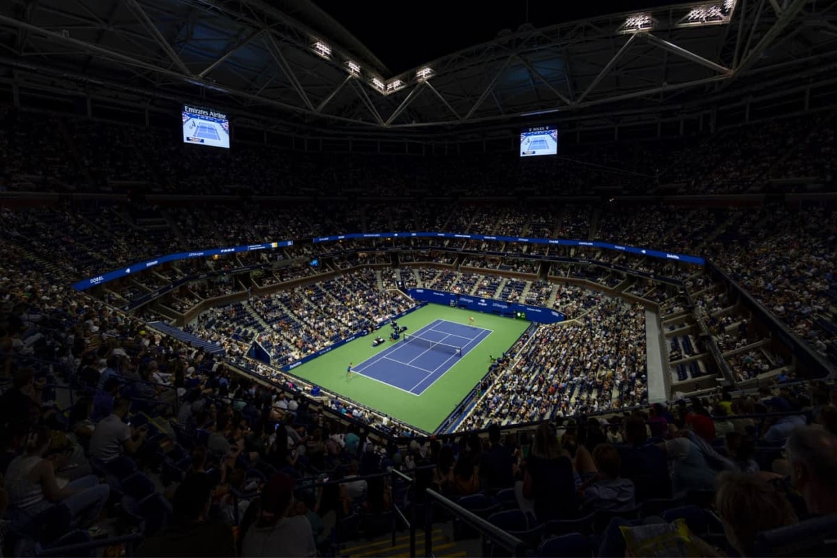Ein voll besetztes Tennisstadion bei Nacht. Auf dem hell erleuchteten blauen Platz läuft gerade ein Spiel. Das Publikum steht um den Platz herum und auf zwei großen Bildschirmen werden Spieldetails angezeigt. Das Dach des Stadions ist teilweise sichtbar.