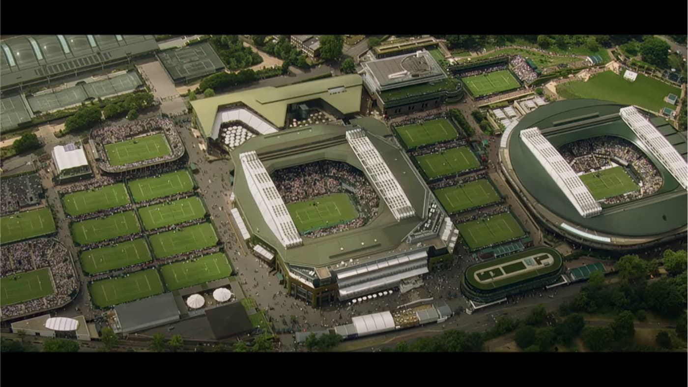 Luftaufnahme eines großen Tenniskomplexes mit mehreren Plätzen, darunter zwei Hauptstadien. Menschenmengen versammeln sich um die Plätze, umgeben von üppigem Grün und Wegen, was ein geschäftiges Sportereignis darstellt.