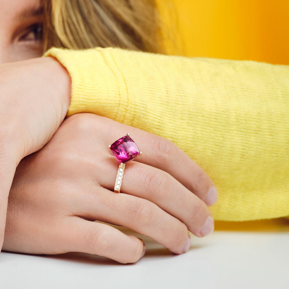 Eine Person in einem gelben Pullover legt ihren Kopf auf ihre verschränkten Arme und präsentiert den eleganten RUBELLITRING von Schullin. Der Ring zeigt einen bezaubernden Rubellit, der in ein mit kleinen Diamanten verziertes Roségoldband eingefasst ist. Der Hintergrund ergänzt die Szene mit seinem kräftigen Gelbton.