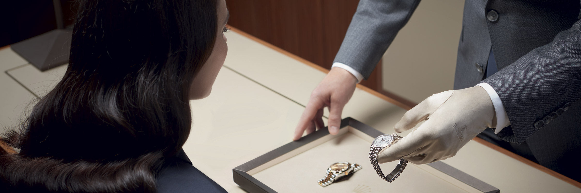 Eine Person in Anzug und Handschuhen präsentiert einem sitzenden Kunden eine Luxusuhr von einem Tablett. Auf dem Tablett liegt außerdem eine weitere Uhr, und die Umgebung scheint eine Luxusboutique oder ein Juweliergeschäft zu sein.