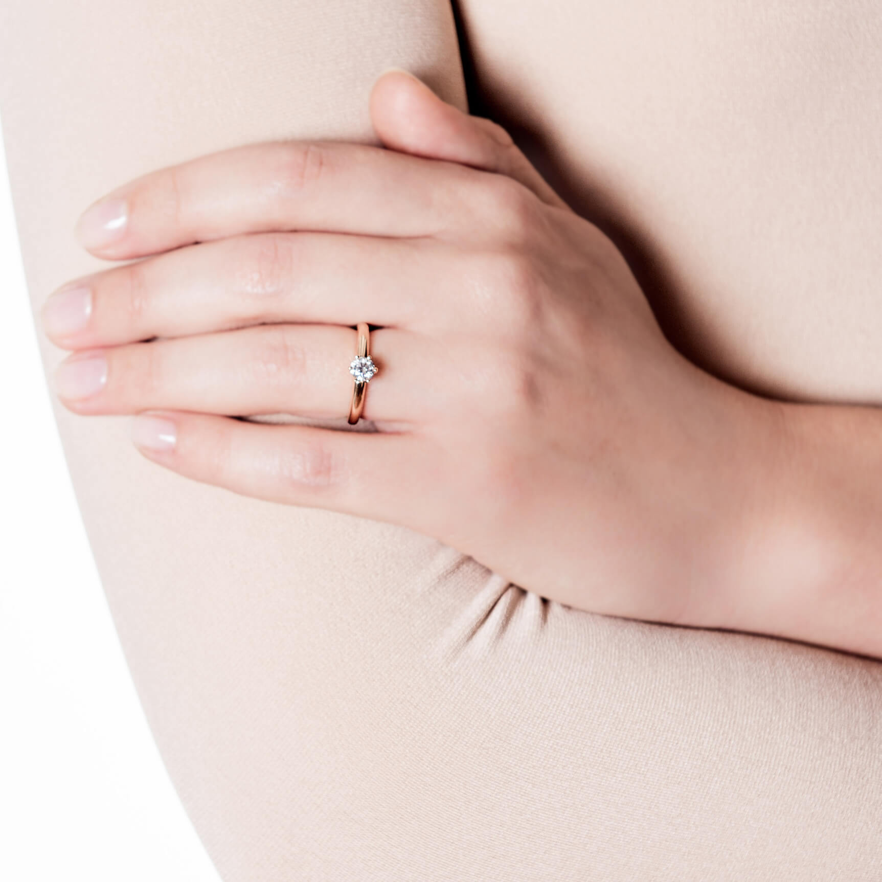 Eine Nahaufnahme der Hand einer Person, die mit dem PRINCESS SOLITÄR 0,50 CARAT Ring aus 18 Karat Roségold von Schullin geschmückt ist, der mit einem einzelnen runden Diamanten versehen ist. Die Hand ruht sanft auf dem gegenüberliegenden Arm, der in beigen Stoff gehüllt ist.