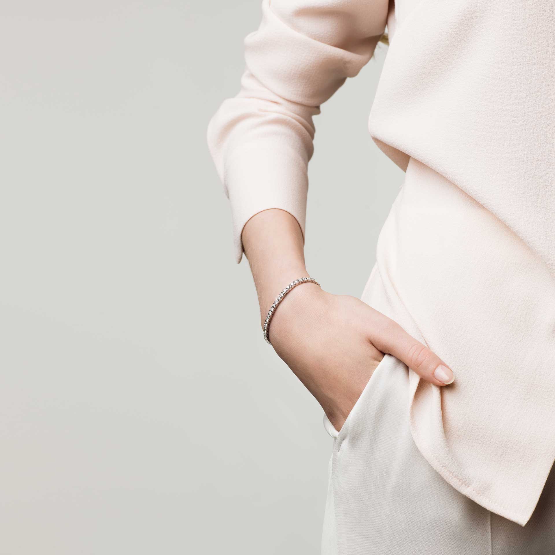 Eine Person trägt ein hellrosa Langarmshirt und eine weiße Hose und hat eine Hand in der Tasche. Der andere Arm hängt entspannt neben ihr und präsentiert ein zartes Schullin Tennisarmband 0,75 Karat Weißgold vor einem schlichten, gedeckten Hintergrund.