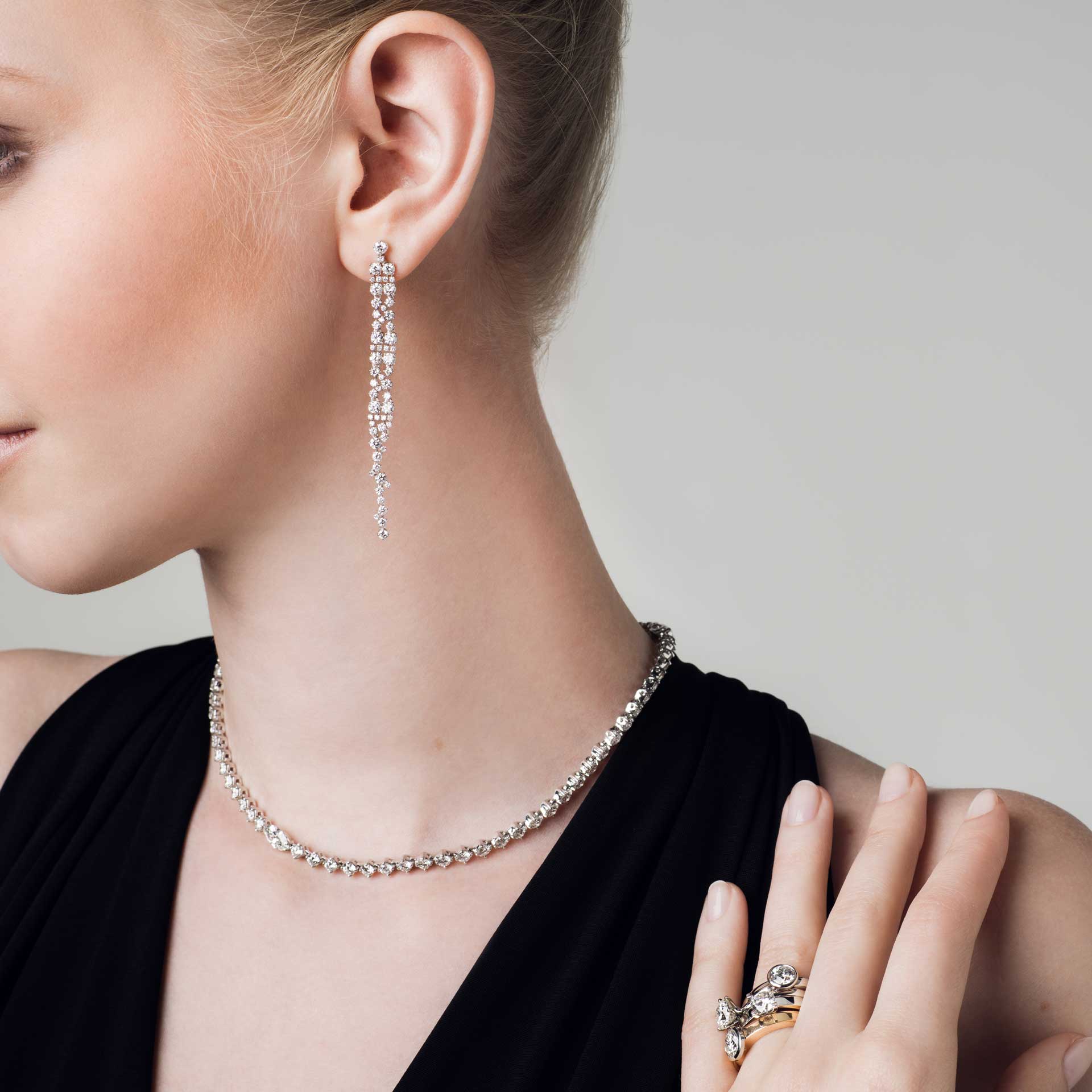 Eine Person im schwarzen Outfit präsentiert eleganten Schmuck, darunter Schullins DIAMANT OHRRINGE mit Brillant-Schliff, eine zarte Halskette aus 18 Karat Weißgold und einen mit Edelsteinen verzierten Ring. Der Fokus liegt auf der Brillanz des Schmucks vor einem neutralen Hintergrund.