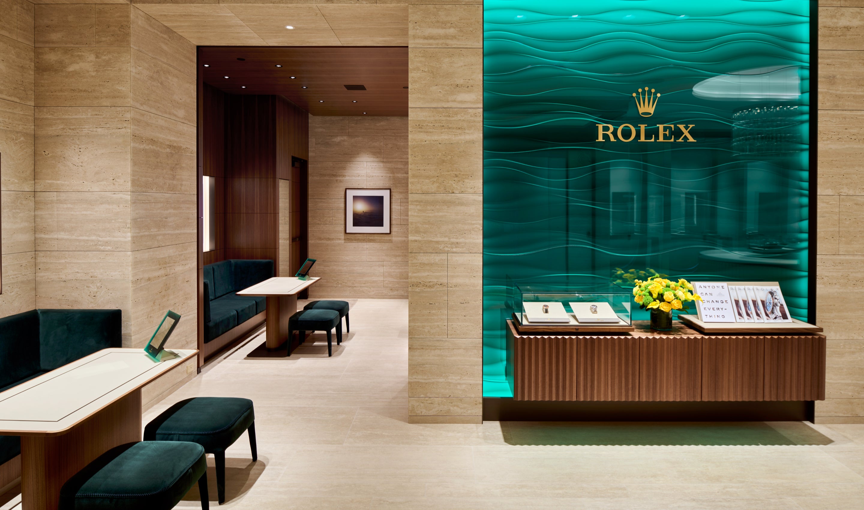 Luxuriöses Interieur einer Uhrenboutique mit Rolex-Ausstellung im türkisfarbenen Wellendesign und goldenem Logo. Elegante Sitzgelegenheiten mit Holztischen säumen die beigen Steinwände und schaffen eine gehobene Atmosphäre.
