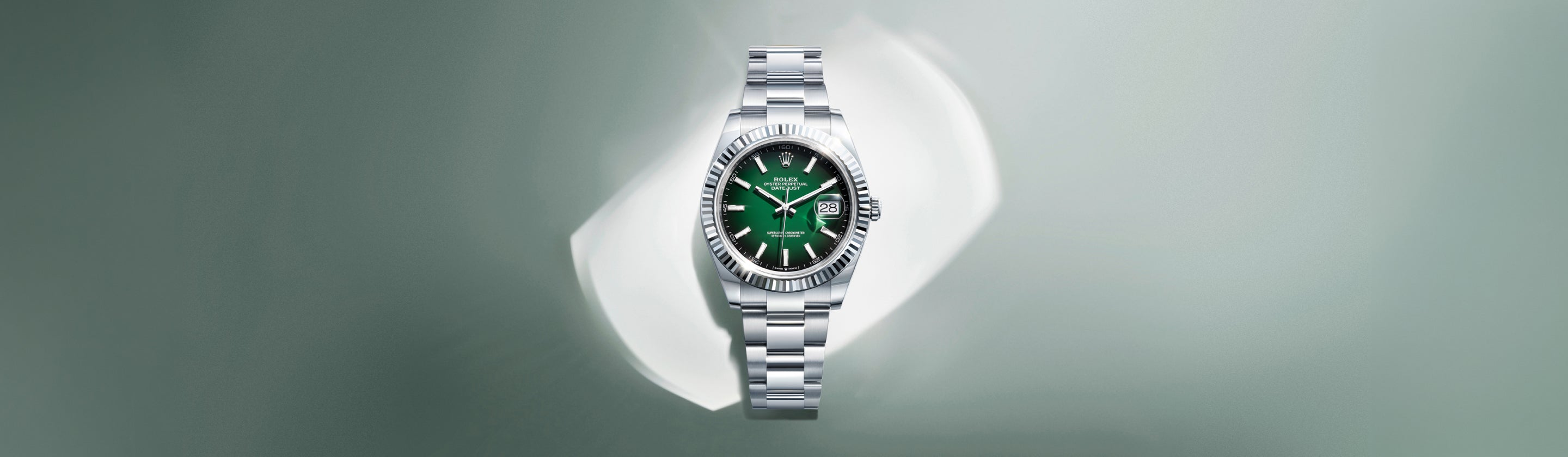 Eine silberne Armbanduhr mit grünem Zifferblatt und Datumsanzeige ist vor einem weichen, hellgrauen Hintergrund mit Farbverlauf zentriert. Die Uhr hat ein Metallgliederarmband und eine geriffelte Lünette.