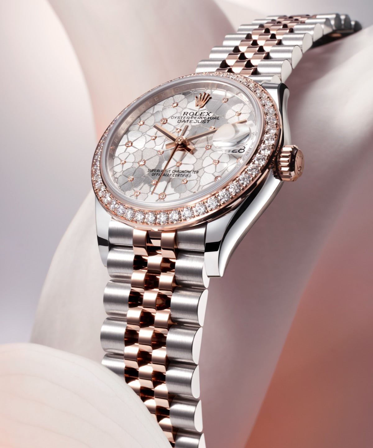 Eine Nahaufnahme einer Luxusuhr mit einem Armband aus Silber und Roségold. Das Zifferblatt ist mit einem Blumenmuster verziert, Diamantakzente um die Lünette herum und der Markenname ist zu sehen. Er wird elegant auf einem weichen, abstrakten Hintergrund dargestellt.