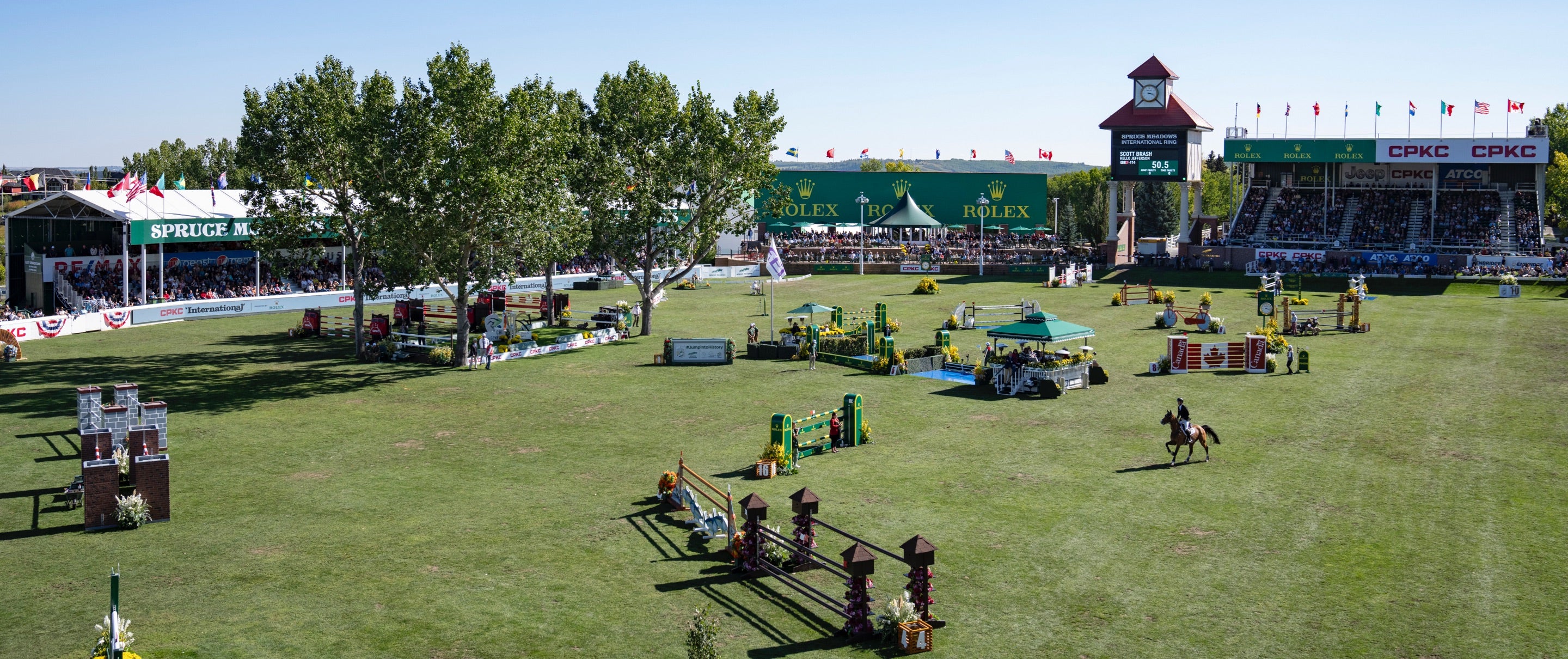 Eine weite Sicht auf einen Springwettbewerb in Spruce Meadows. Die Arena ist von Zuschauern unter Zelten und Sitzplätzen umgeben. Auf dem Grasfeld sind verschiedene farbenfrohe Hindernisse aufgebaut und man sieht einen Reiter auf einem Pferd in Aktion.