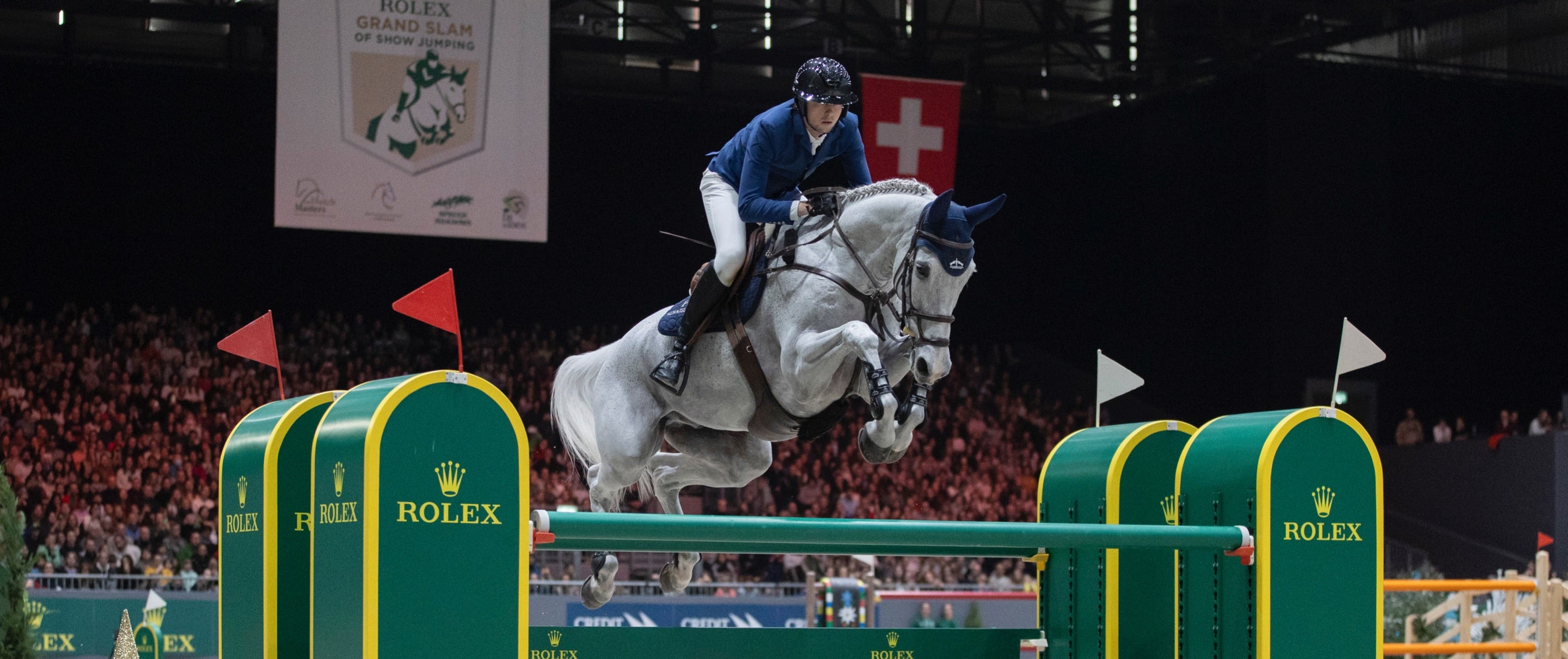 Ein Reiter in blauem Outfit springt bei einem Indoor-Springturnier auf einem weißen Pferd über ein grün-gelbes Hindernis mit Rolex-Logo. Im Hintergrund sind das dicht gefüllte Publikum und ein Veranstaltungsbanner zu sehen.