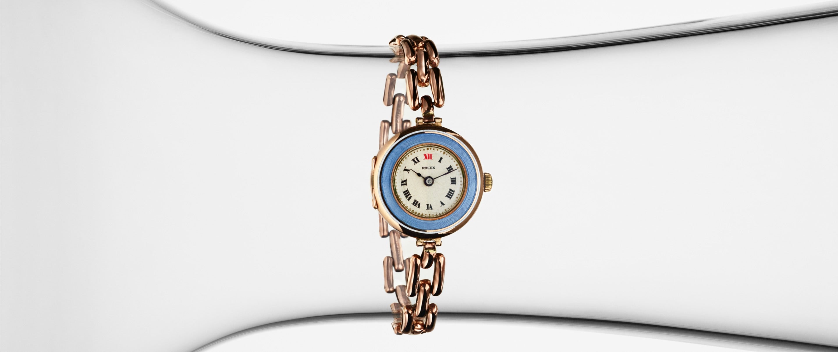 Eine goldene Armbanduhr mit blauer Lünette und Gliederarmband ist zentriert vor einem weißen Hintergrund. Das Zifferblatt zeigt römische Ziffern und feine Zeiger. Das elegante Design der Uhr zeichnet sich durch rote Stundenmarkierungen bei 3, 6, 9 und 12 aus.