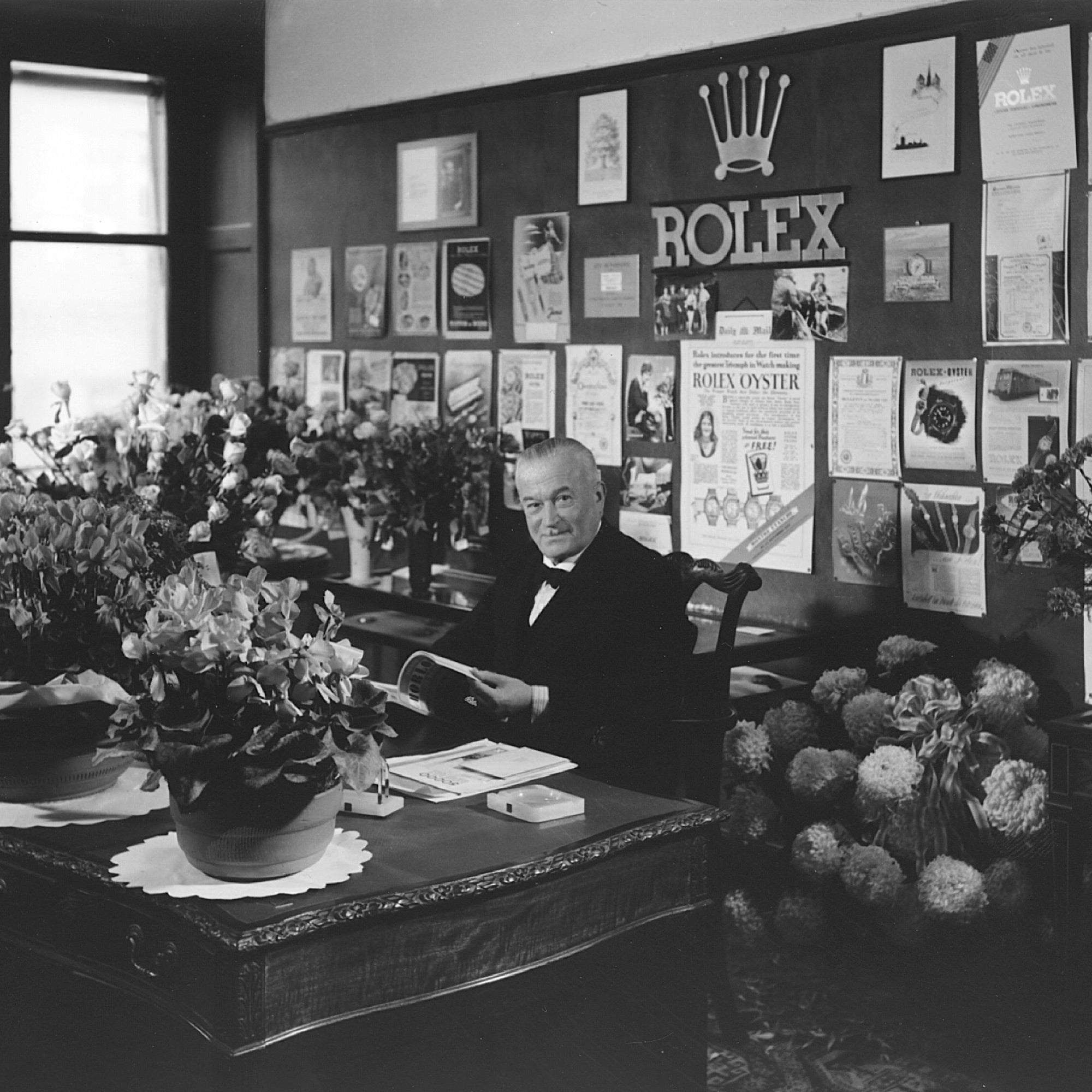 Ein Mann sitzt an einem Holzschreibtisch in einem Büro, das mit verschiedenen Rolex-Werbungen und einem großen Rolex-Kronenlogo an der Wand dekoriert ist. Der Schreibtisch ist mit Blumenarrangements geschmückt und der Mann hält ein Buch in der Hand. Das Zimmer ist mit Vintage-Möbeln ausgestattet.