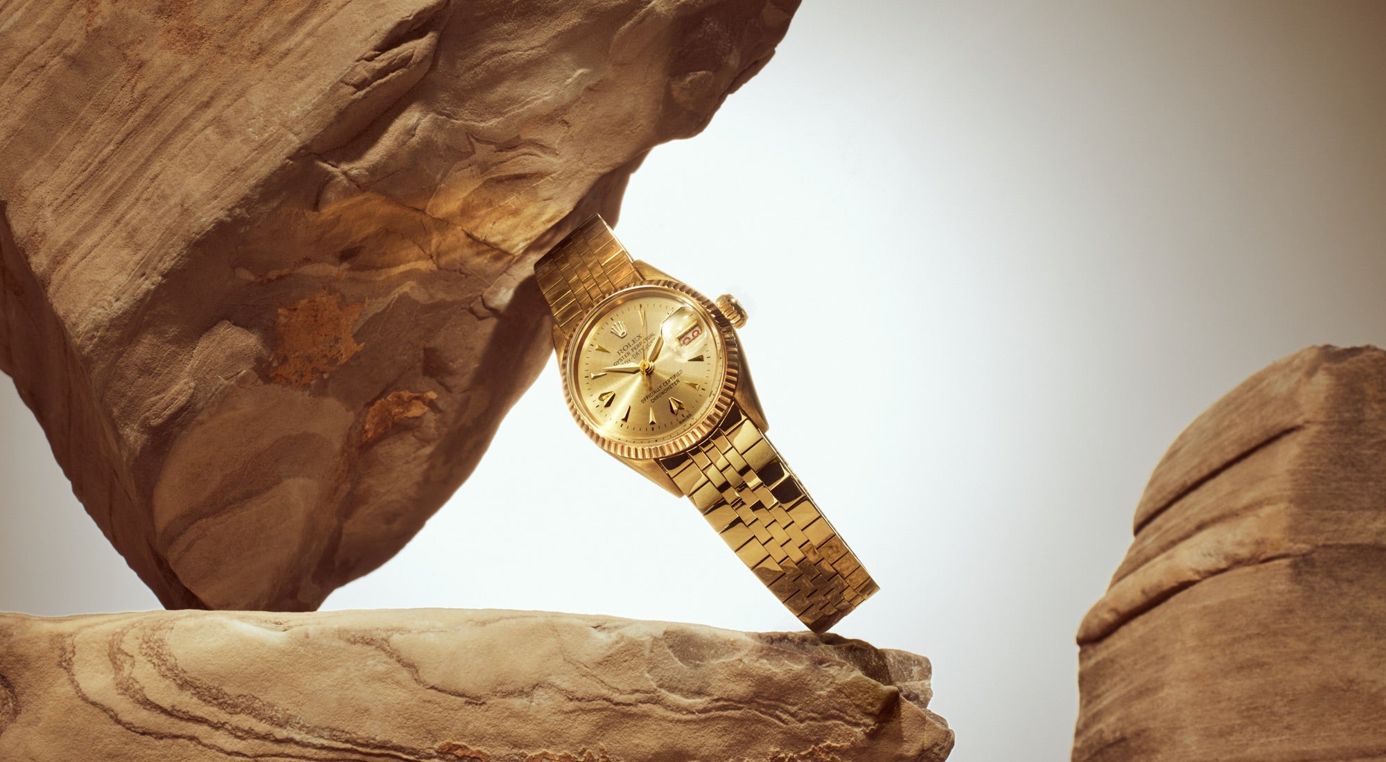 Eine goldene Armbanduhr mit strukturiertem Armband ist kunstvoll zwischen zwei großen, gestapelten Steinen vor einem neutralen Hintergrund platziert und ergibt so ein elegantes und luxuriöses Display.