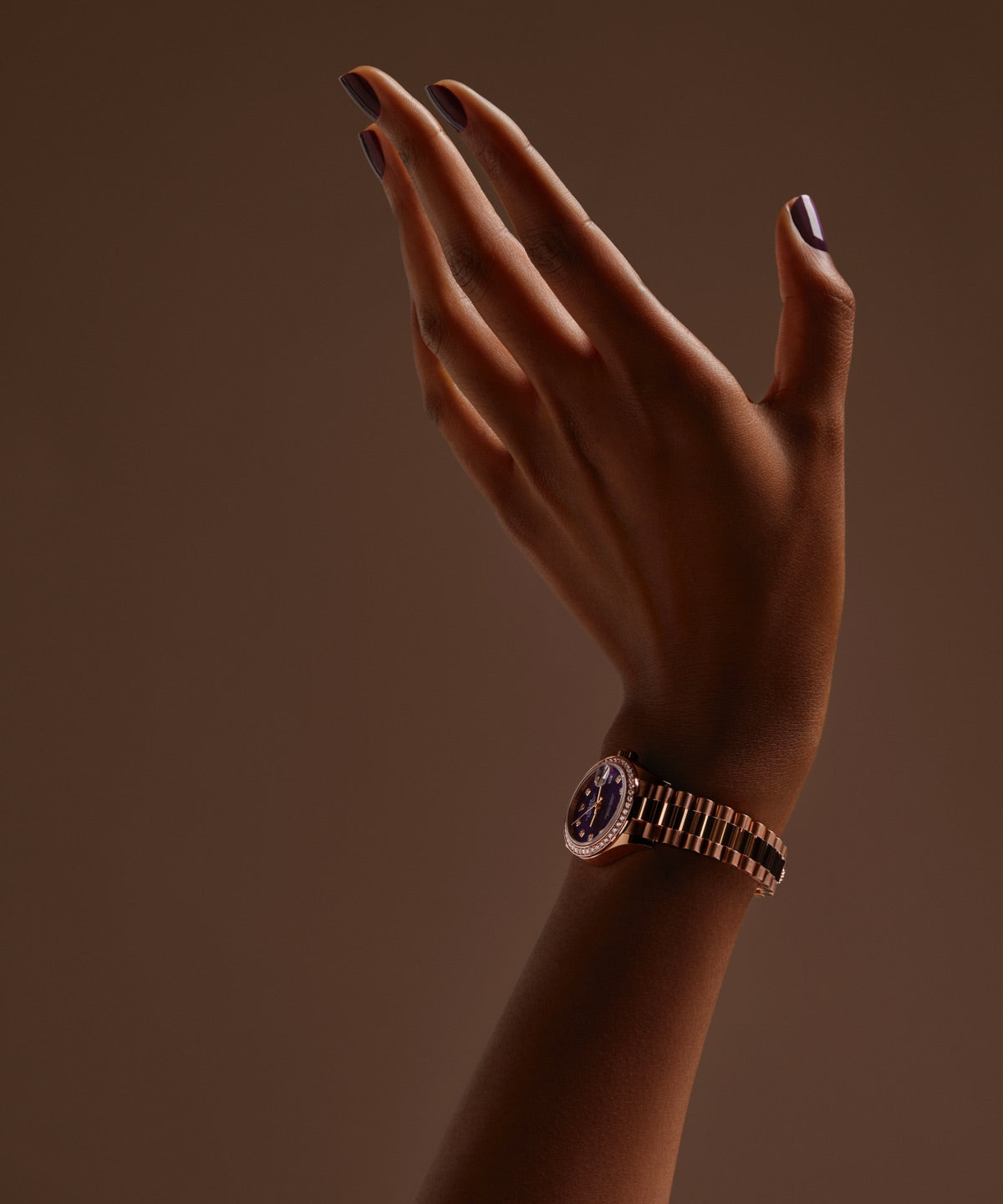Eine Hand mit dunkelviolettem Nagellack posiert elegant vor einem braunen Hintergrund. Das Handgelenk ziert eine roségoldene Uhr mit violettem Ziffernblatt, die der minimalistischen Komposition einen Hauch von Raffinesse verleiht.