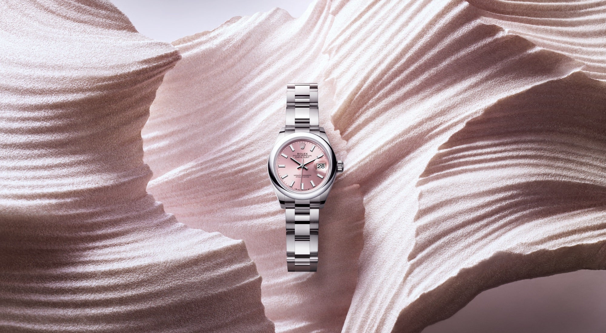 Eine silberne Armbanduhr mit Metallarmband und rosa Zifferblatt steht vor einem strukturierten, fließenden, weißen Hintergrund, der an Stoff oder eine Papierskulptur erinnert. Die Zeiger zeigen auf 10:10 und ihr Design kontrastiert mit dem welligen Hintergrund.