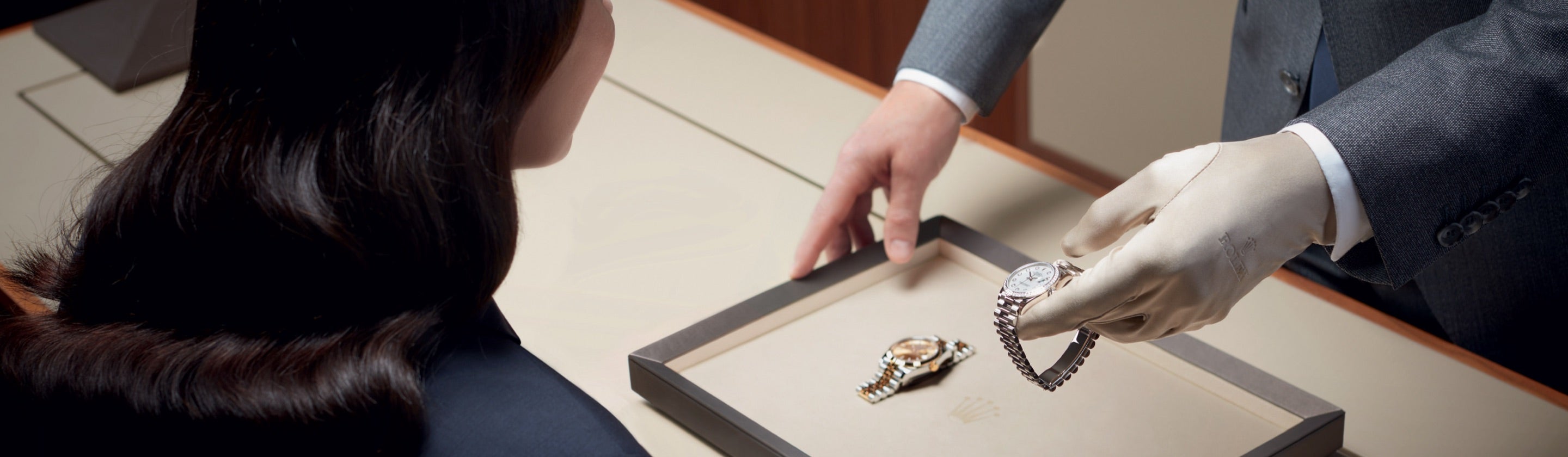 Eine Person im Anzug präsentiert einer anderen Person an einem Tisch eine Luxusuhr. Die Uhr liegt in einer eleganten Box, eine weitere Uhr ist auf dem Tisch zu sehen. Die Person, die die Uhr präsentiert, trägt weiße Handschuhe.