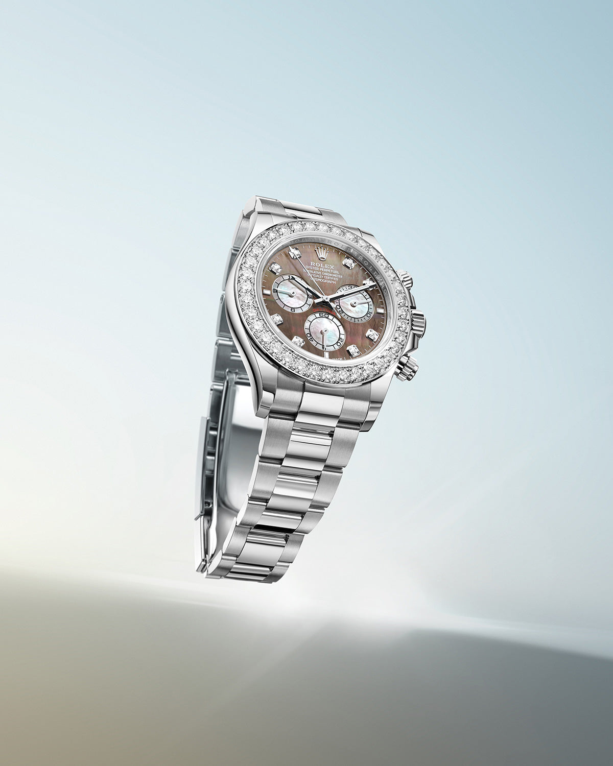 Eine luxuriöse silberne Armbanduhr mit diamantbesetzter Lünette und metallisch braunem Zifferblatt mit mehreren Hilfszifferblättern. Das Armband ist ebenfalls silberfarben und kontrastiert mit einem Hintergrund in Blau- und Beigetönen.
