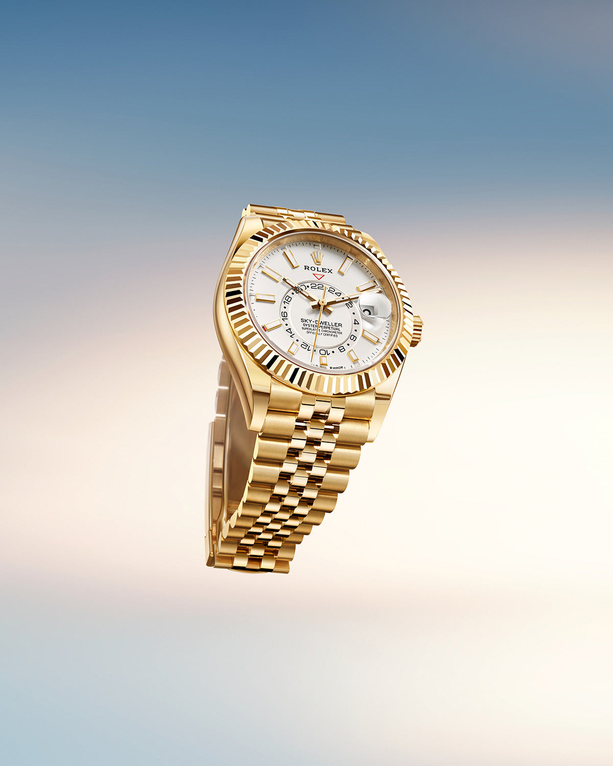 Goldene Armbanduhr mit weißem Zifferblatt und aufwendigen Details auf der Vorderseite sowie einem Gliederarmband. Sie ist auf einem Hintergrund mit Farbverlauf von Hellbeige zu Zartblau abgesetzt.