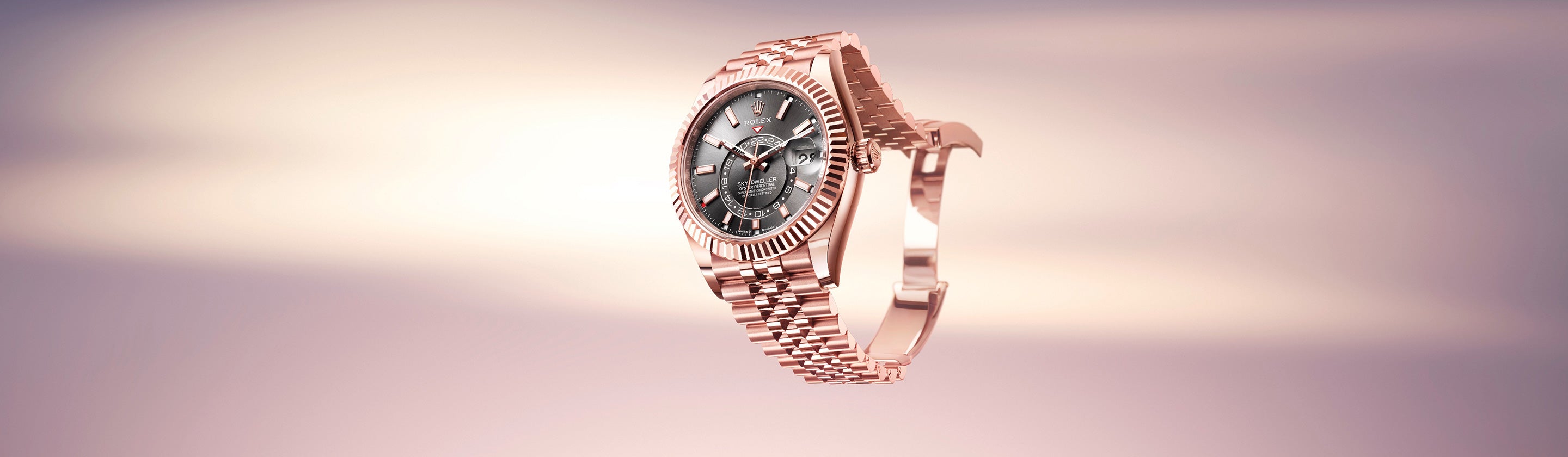 Eine Armbanduhr aus Roségold mit schwarzem Zifferblatt und aufwendigen Details schwebt vor einem sanft verschwommenen Hintergrund mit Farbverlauf in Rosa und Beige. Die Uhr verfügt über ein Gliederarmband und eine gekerbte Lünette.