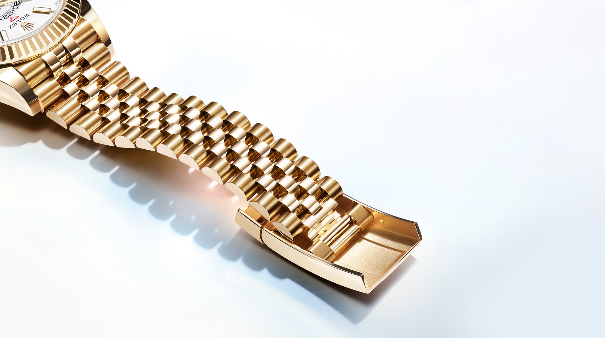 Nahaufnahme einer goldenen Armbanduhr mit Fokus auf dem Armband mit einem polierten, ineinandergreifenden Design. Das Zifferblatt ist teilweise sichtbar und zeigt ein weißes Zifferblatt mit einem einfachen Design vor einem weichen weißen Hintergrund.