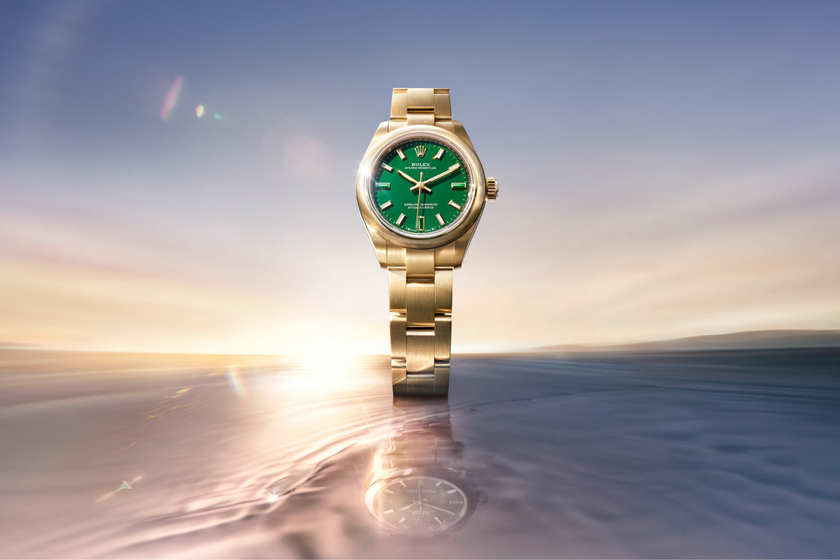 Eine goldene Armbanduhr mit grünem Ziffernblatt steht in der Mitte vor einem weichen, verträumten Hintergrund aus Wasser und einem glühenden Sonnenuntergang, unter dem sich die Uhr spiegelt.