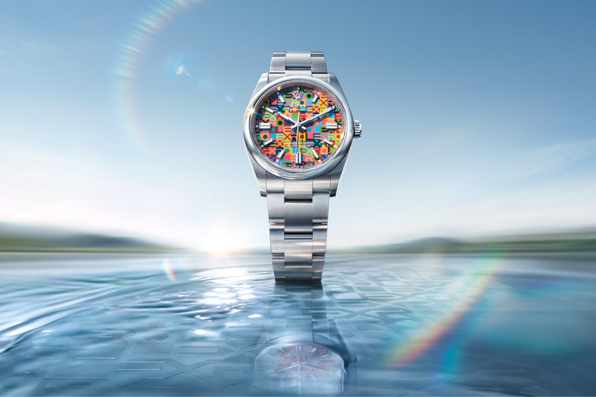Eine Armbanduhr aus Edelstahl mit einem mehrfarbigen, geometrisch gemusterten Zifferblatt steht aufrecht auf einer spiegelnden Wasseroberfläche, mit einer verschwommenen Landschaft und regenbogenartigen Lichteffekten im Hintergrund.
