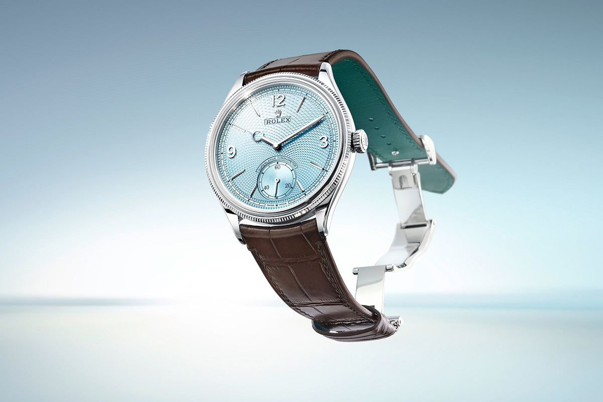 Eine silberne Armbanduhr mit blauem Zifferblatt und braunem Lederarmband steht diagonal vor einem hellblauen Hintergrund mit Farbverlauf. Das Zifferblatt verfügt über Ziffern und zwei Hilfszifferblätter.