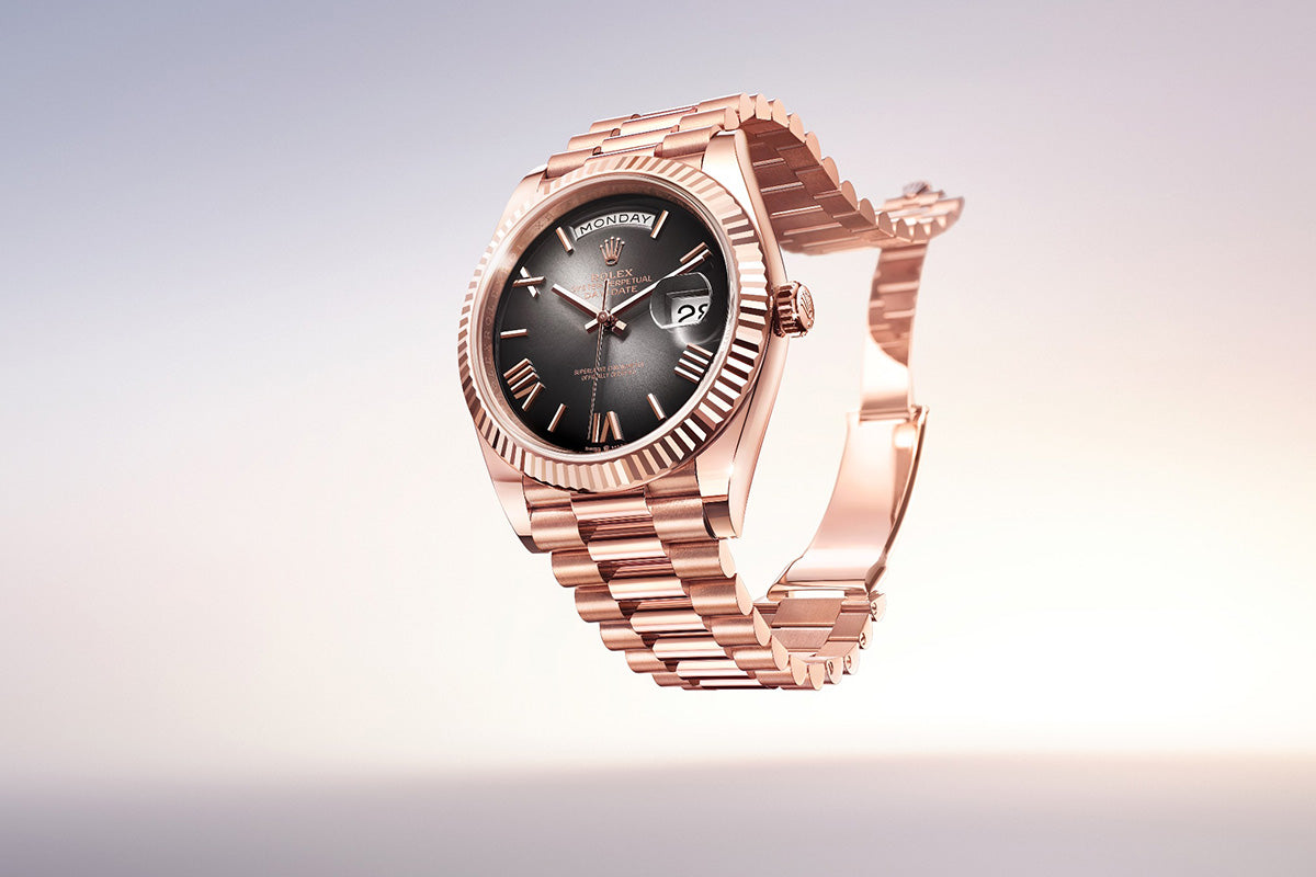 Eine luxuriöse Armbanduhr aus Roségold mit schwarzem Zifferblatt und Tag-/Datumsanzeige. Die Uhr verfügt über eine geriffelte Lünette und ihr Metallarmband ist elegant gestaltet und hebt sich von einem Hintergrund mit Farbverlauf in sanften Weiß- bis Pfirsichtönen ab.