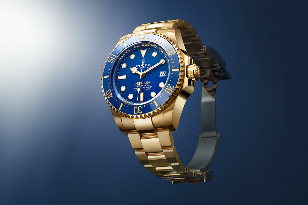 Eine luxuriöse goldene Uhr mit blauem Zifferblatt und Lünette, Datumsanzeige und Leuchtmarkierungen. Die Uhr ist auf einem dunkelblauen Hintergrund angebracht und ein Spotlight hebt die polierte Oberfläche hervor.