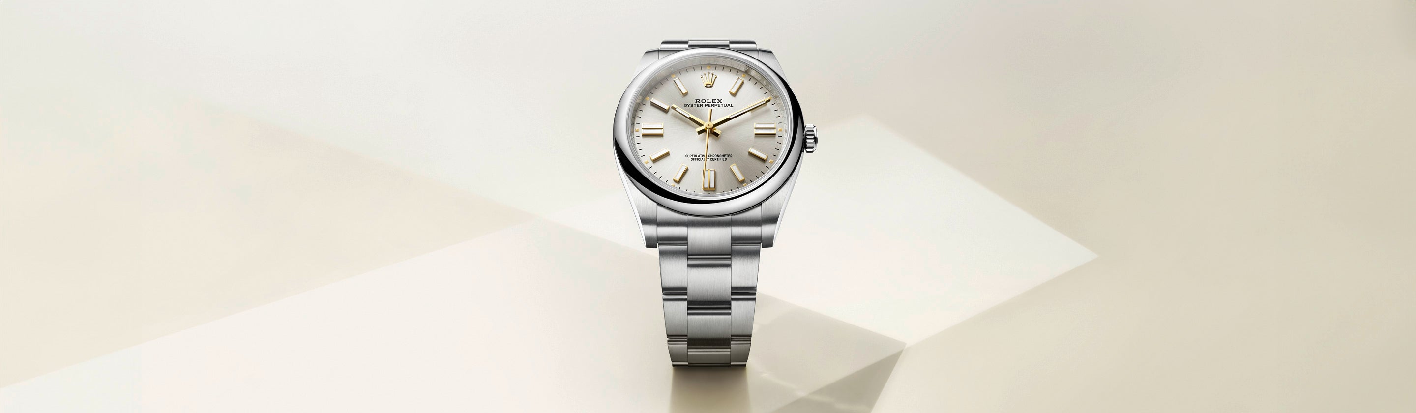 Eine silberne Armbanduhr mit Edelstahlarmband und weißem Zifferblatt wird vor einem weichen, cremefarbenen geometrischen Hintergrund präsentiert. Die Uhr verfügt über goldene Stundenmarkierungen und Zeiger.