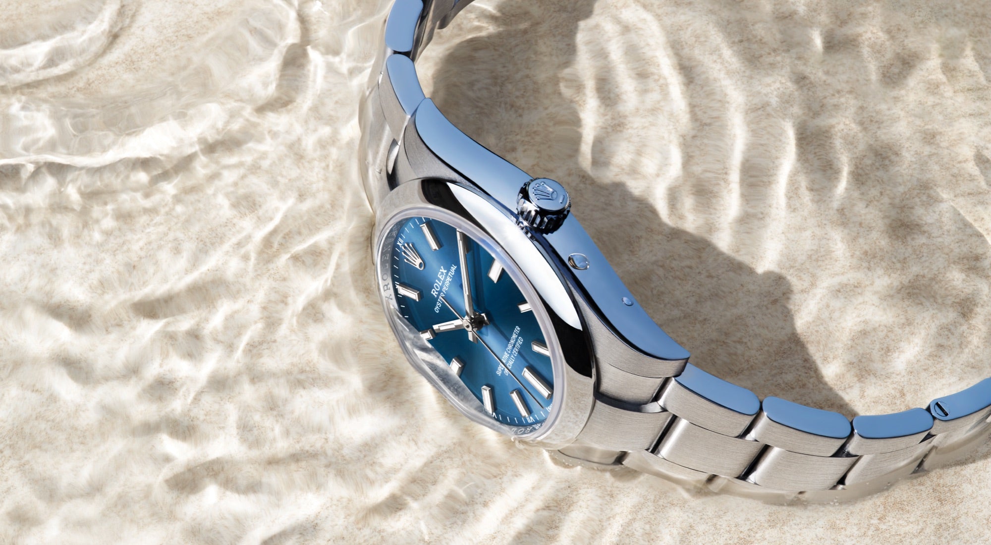 Eine silberne Armbanduhr mit blauem Zifferblatt und silbernen Stundenmarkierungen wird auf einer strukturierten, sandigen Oberfläche gezeigt. Das Licht erzeugt Reflexionen und Schatten auf dem Metallarmband und hebt das schlanke Design und die polierte Oberfläche der Uhr hervor.