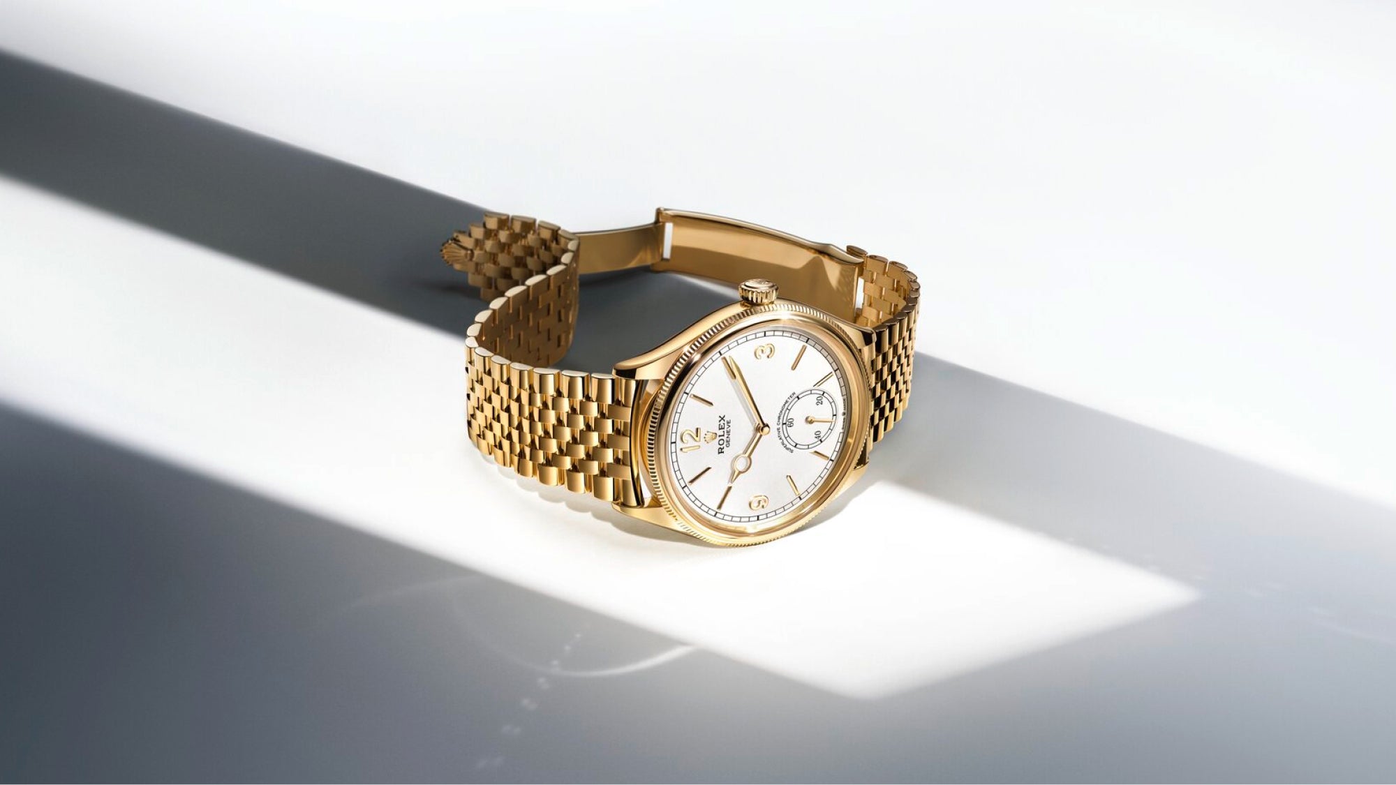 Eine goldene Armbanduhr mit weißem Zifferblatt und römischen Ziffern liegt auf einer ebenen Fläche, die teilweise von einem diagonalen Lichtstrahl beleuchtet wird. Das goldene Armband ist geöffnet und die Uhr wirft einen weichen Schatten.