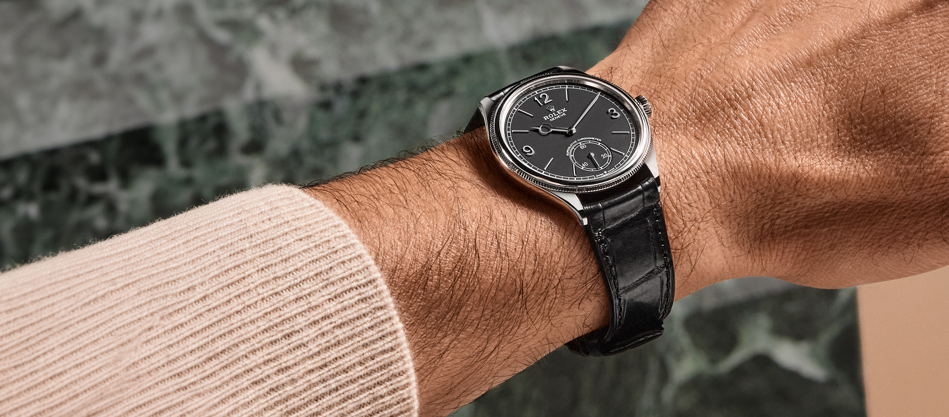 Eine Nahaufnahme des Handgelenks einer Person, die eine Armbanduhr mit schwarzem Lederarmband, schwarzem Zifferblatt und silbernem Gehäuse trägt. Die Person trägt einen hellbeigen Pullover. Der Hintergrund ist unscharf mit Grün- und Beigetönen.