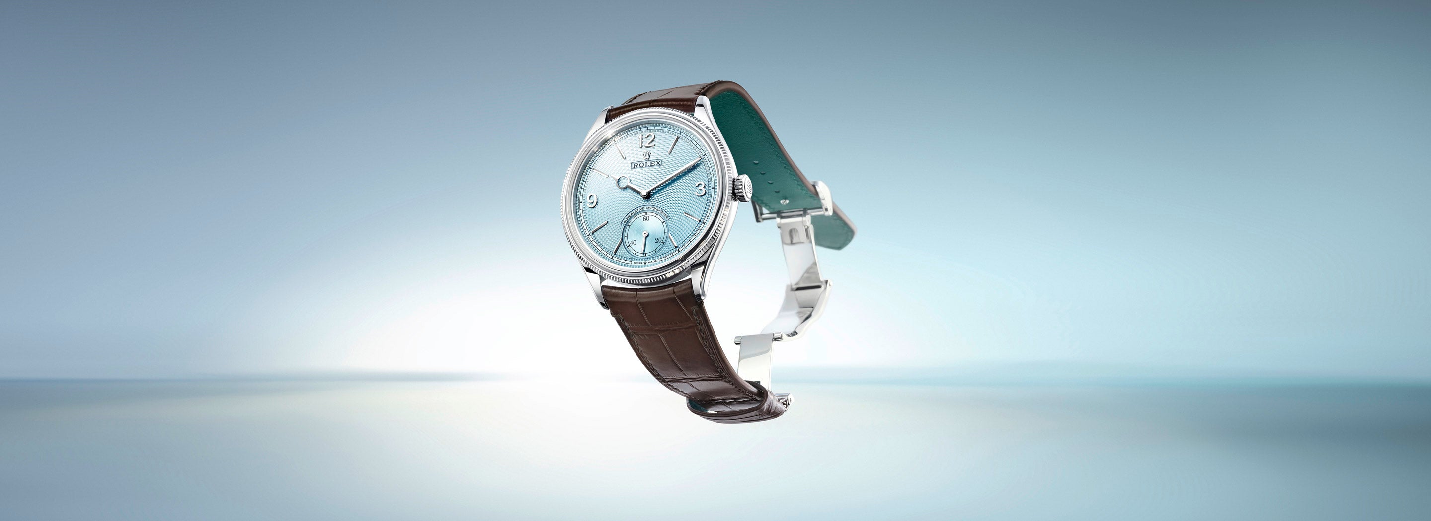 Eine Luxusarmbanduhr mit türkisfarbenem Zifferblatt und silbernen Akzenten und Lederarmband wird vor einem Hintergrund mit Farbverlauf in Blau präsentiert. Die Uhr verfügt über ein kleines Hilfszifferblatt und elegante Stundenmarkierungen.