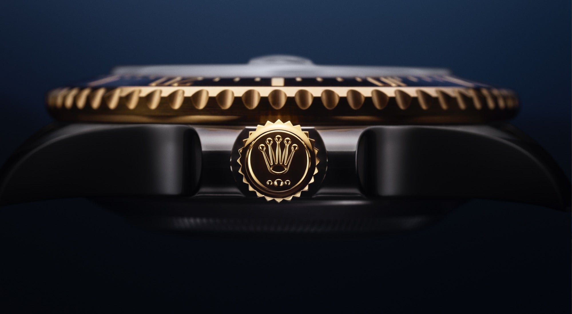 Nahaufnahme der Krone einer Luxusuhr mit einem goldenen Emblem auf einer schwarzen Lünette und einer gezackten Kante auf dem Zifferblatt. Der Hintergrund ist ein dunkelblauer Farbverlauf.