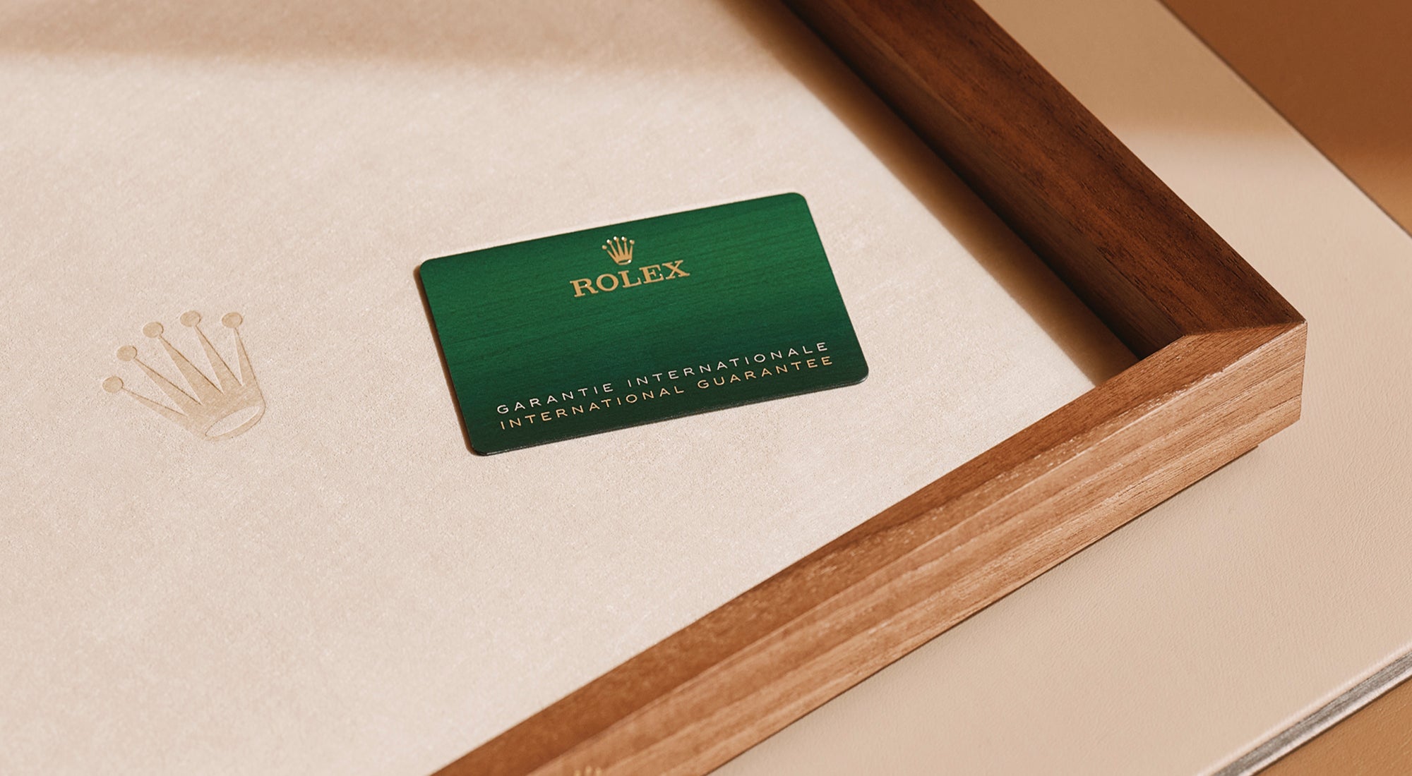 Eine grüne internationale Rolex-Garantiekarte liegt auf einer beigen Stoffoberfläche in einem Holztablett. Links neben der Karte ist ein goldenes Rolex-Kronenlogo auf den Stoff geprägt.