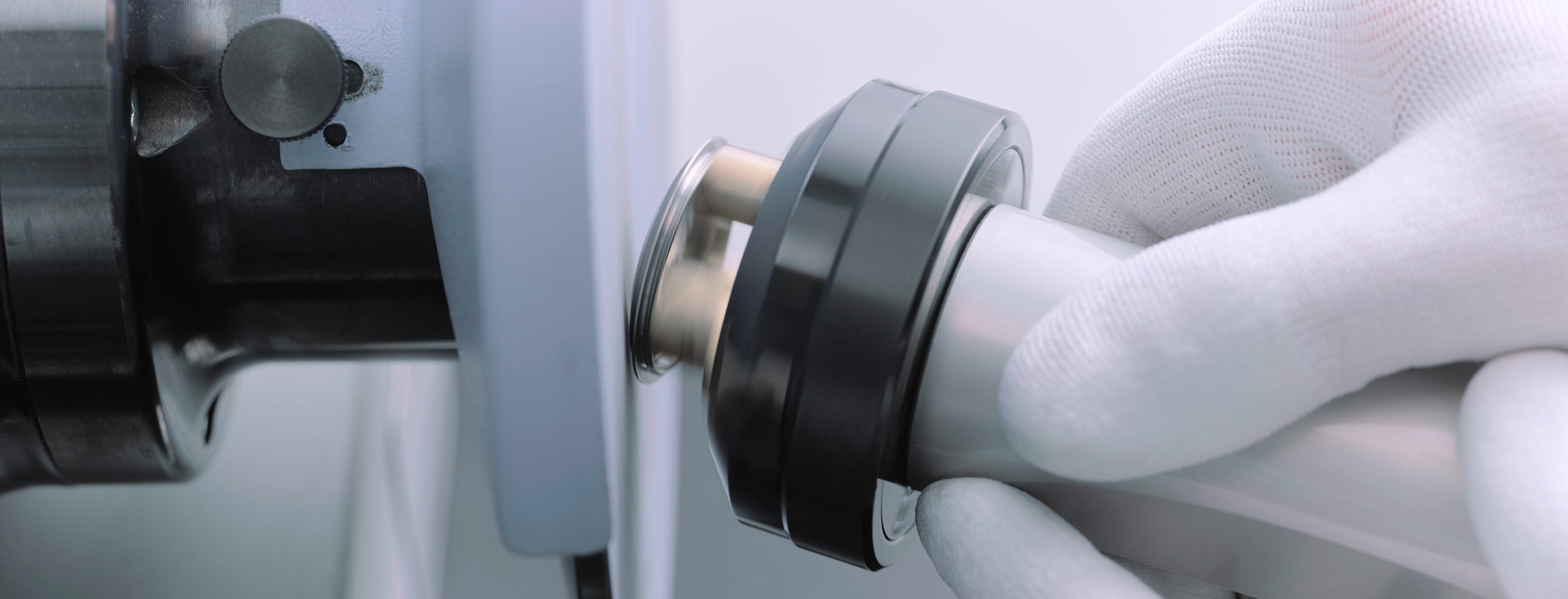 Eine behandschuhte Hand hält in einer Werkstatt ein zylindrisches Werkzeug an eine Schleif- oder Poliermaschine. Die Maschine hat eine metallische Oberfläche und das Werkzeug scheint gerade geschärft oder poliert zu werden.